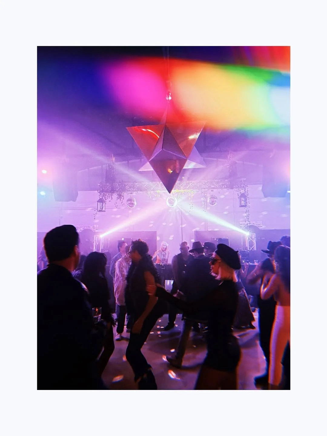 People dancing at a nightclub with colorful lighting, a disco ball, and a geometric hanging decoration.