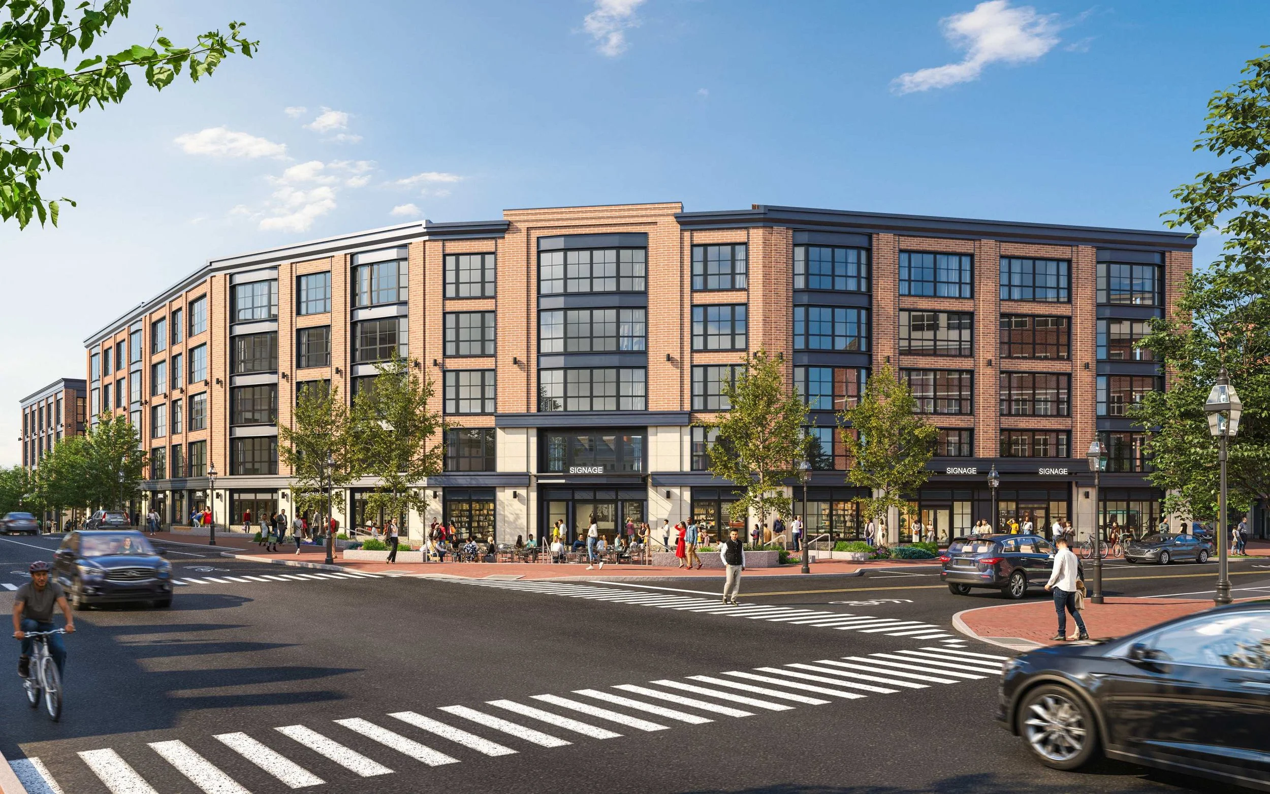 A busy city street scene showing a modern, multi-story brick building with large windows and retail spaces at the ground level. People are walking on the sidewalk, and cars are driving on the street. There are trees and street lamps along the sidewal