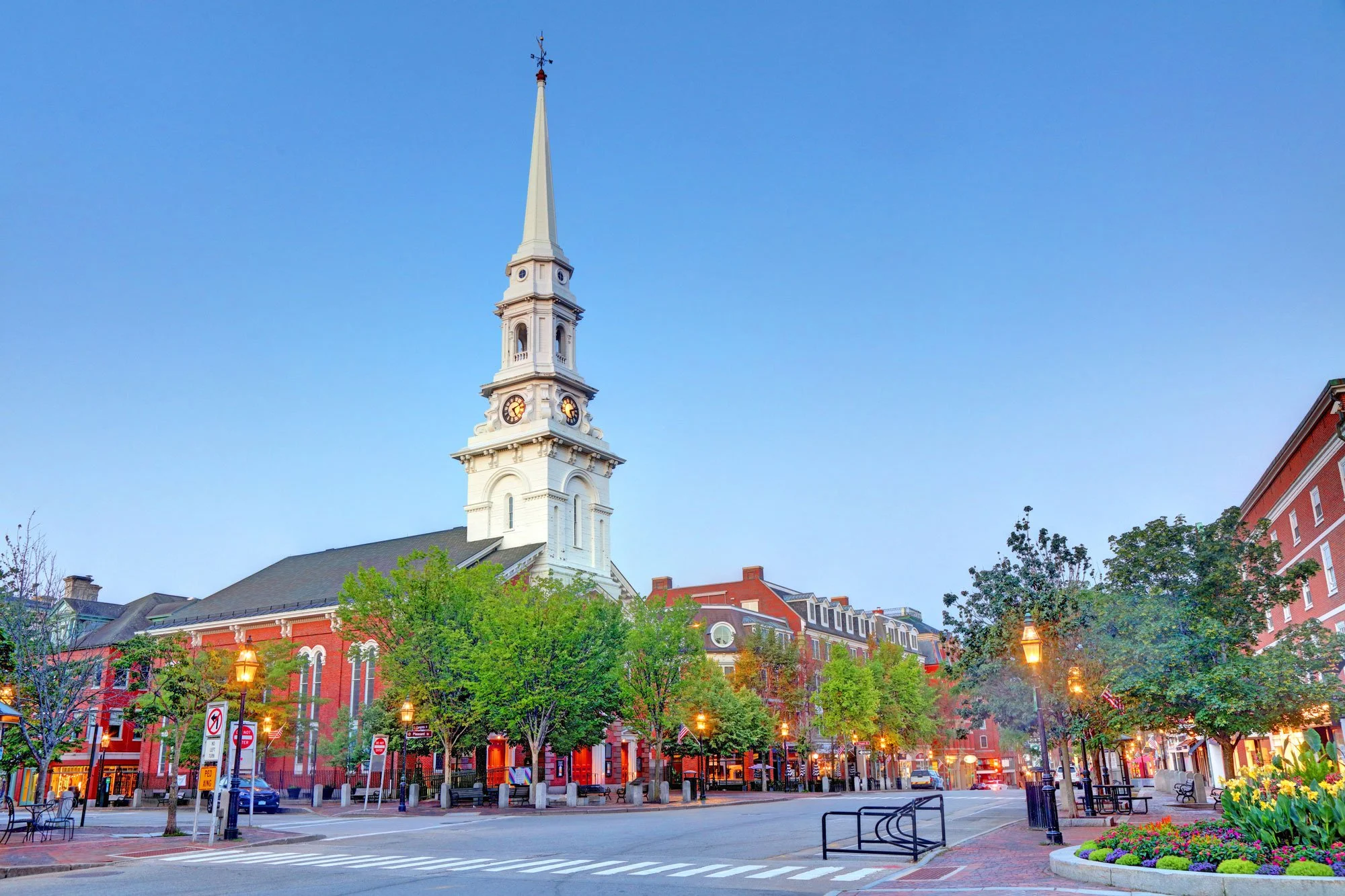 A historic white church with a tall steeple and clock towers, surrounded by a lively city square with trees, street lamps, and colorful buildings in the background during twilight.