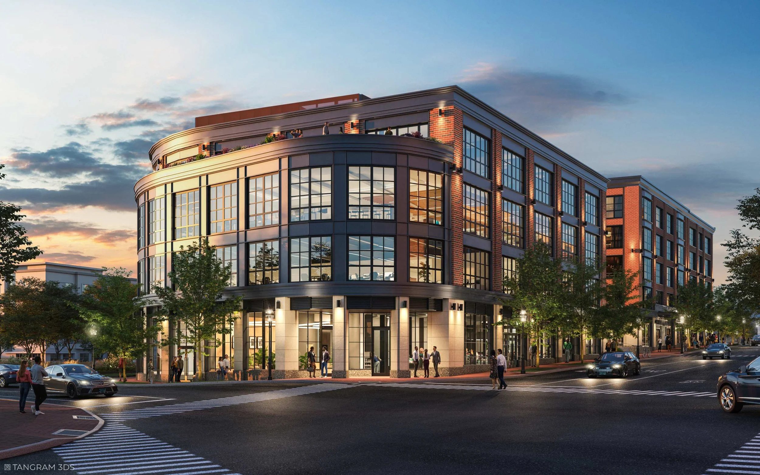 A modern multi-story building with large glass windows, brick accents, and a rounded corner, illuminated at dusk, with people walking and cars parked in front, and trees lining the street.
