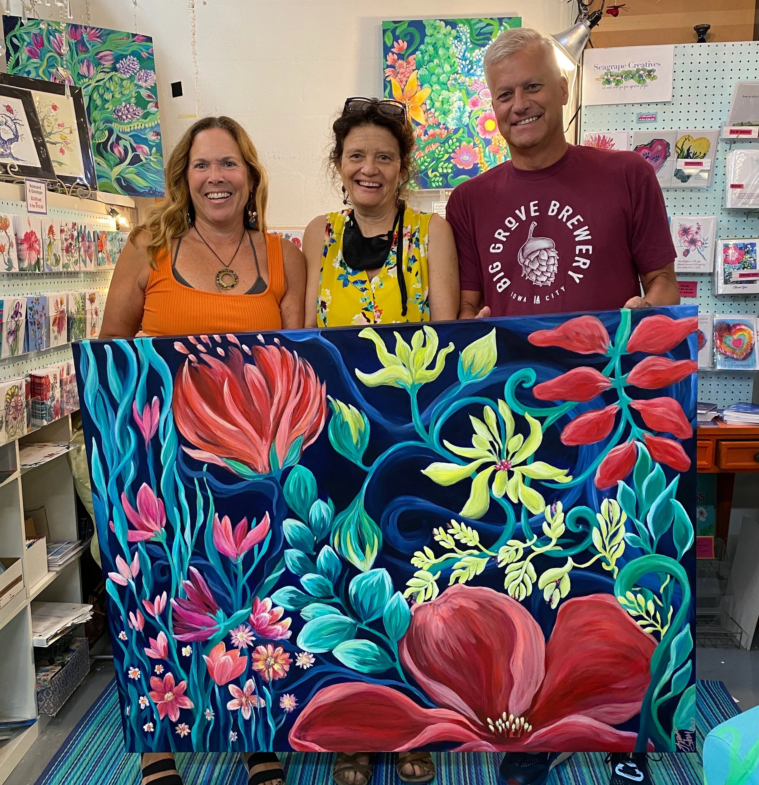 Three people smiling at an art exhibit, with colorful floral paintings on display behind them and a large vibrant floral artwork in front of them.
