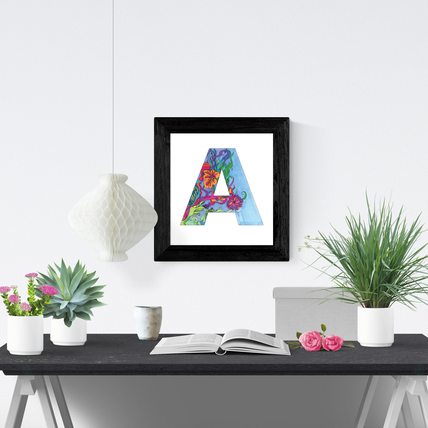 Decorative white paper lantern hanging above a black table with potted plants, an open book, and framed colorful letter 'A' on wall.