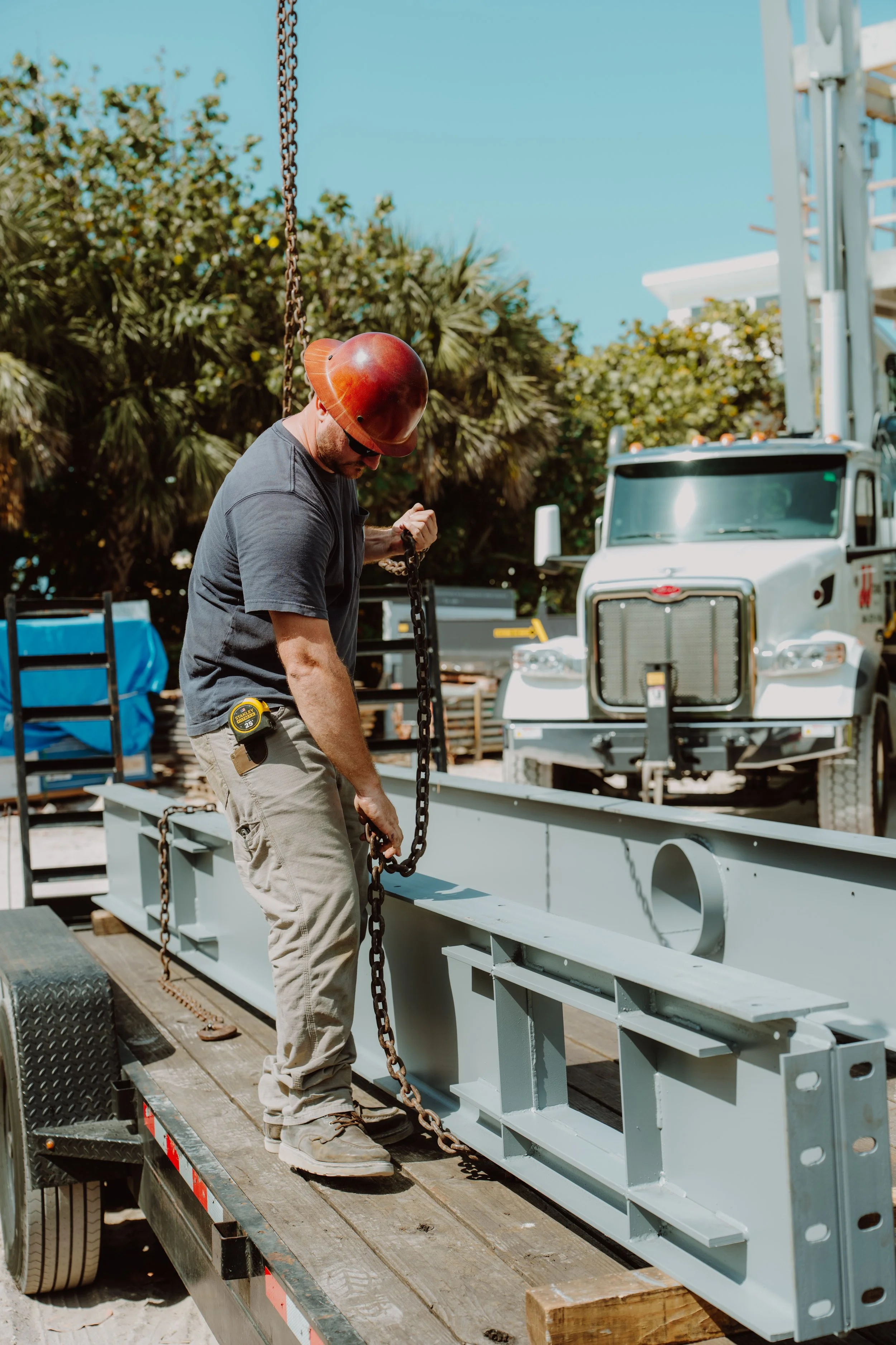 A construction worker wearing a safety helmet and sunglasses is handling a chain on a construction site. A large piece of industrial equipment is on a flatbed trailer, and a tow truck is visible in the background. The scene is outdoors with trees and a clear blue sky.