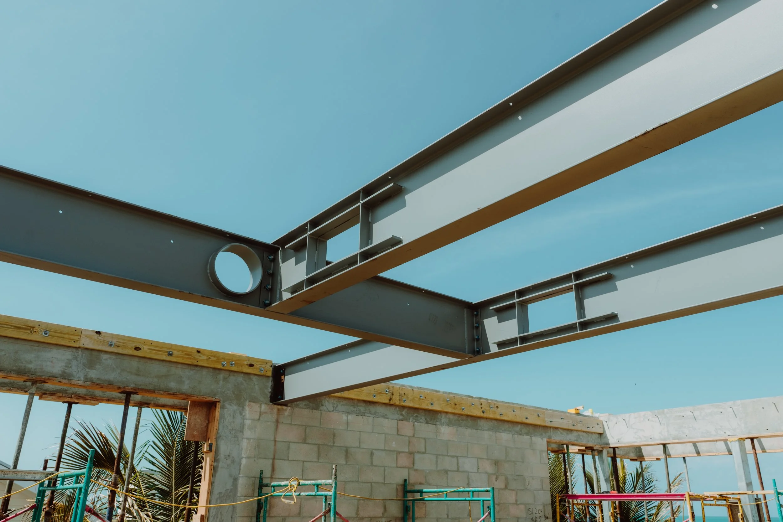Construction site with partially built concrete and metal framing structure, under blue sky, with scattered palm trees in the background.