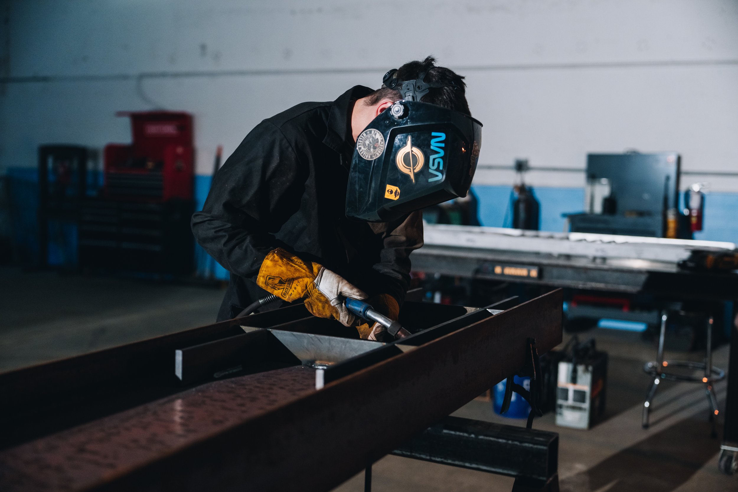Welding professional wearing a protective helmet with stickers, working on a metal piece in a workshop. The workshop has tools, a toolbox, and safety equipment in the background.