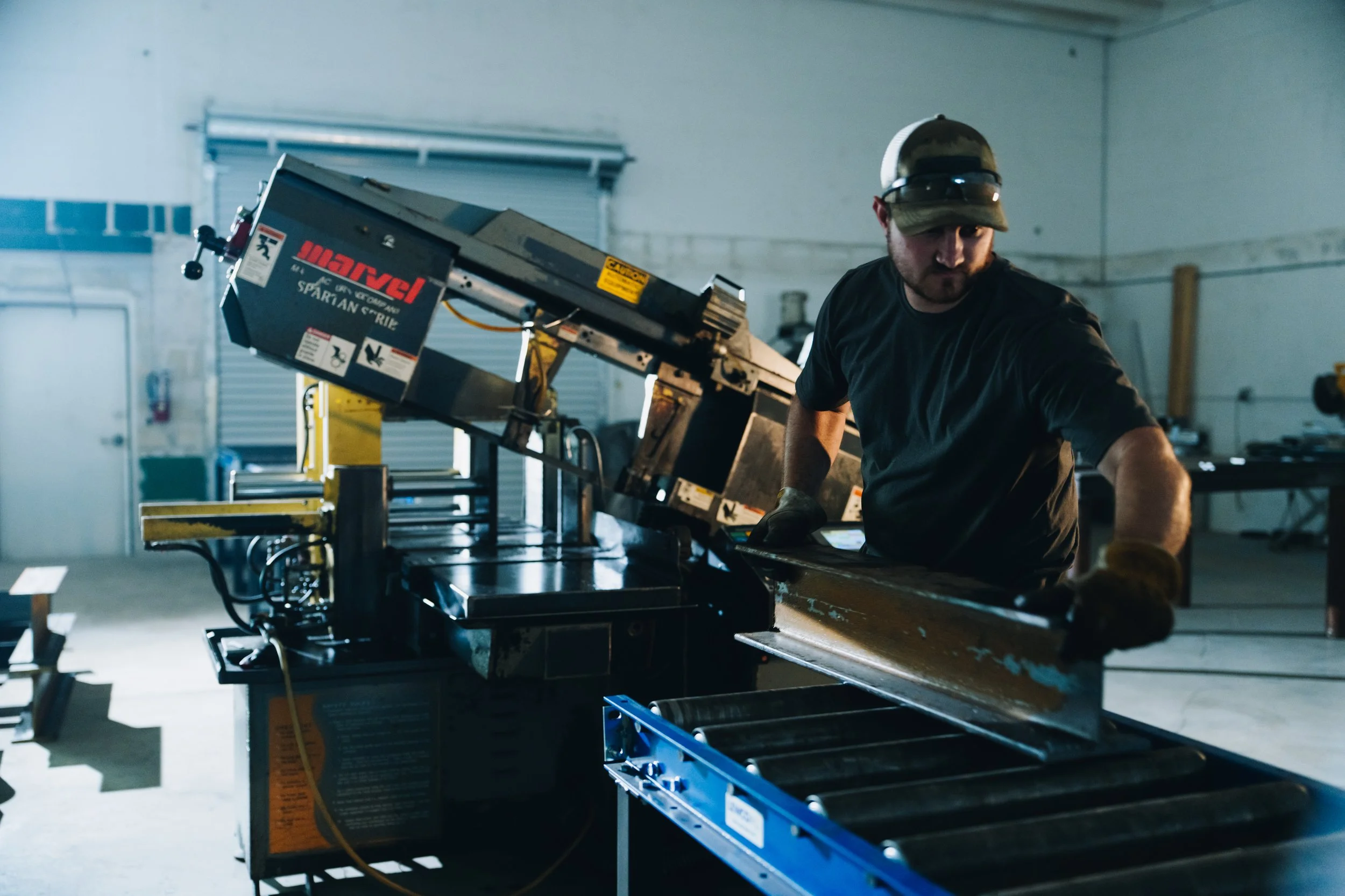 A man with a beard wearing a baseball cap, sunglasses, and a black T-shirt is working with power tools in a garage or workshop. He is near a metal workbench with various equipment on it, and a large black machine labeled "Marvel" is positioned above the work surface.