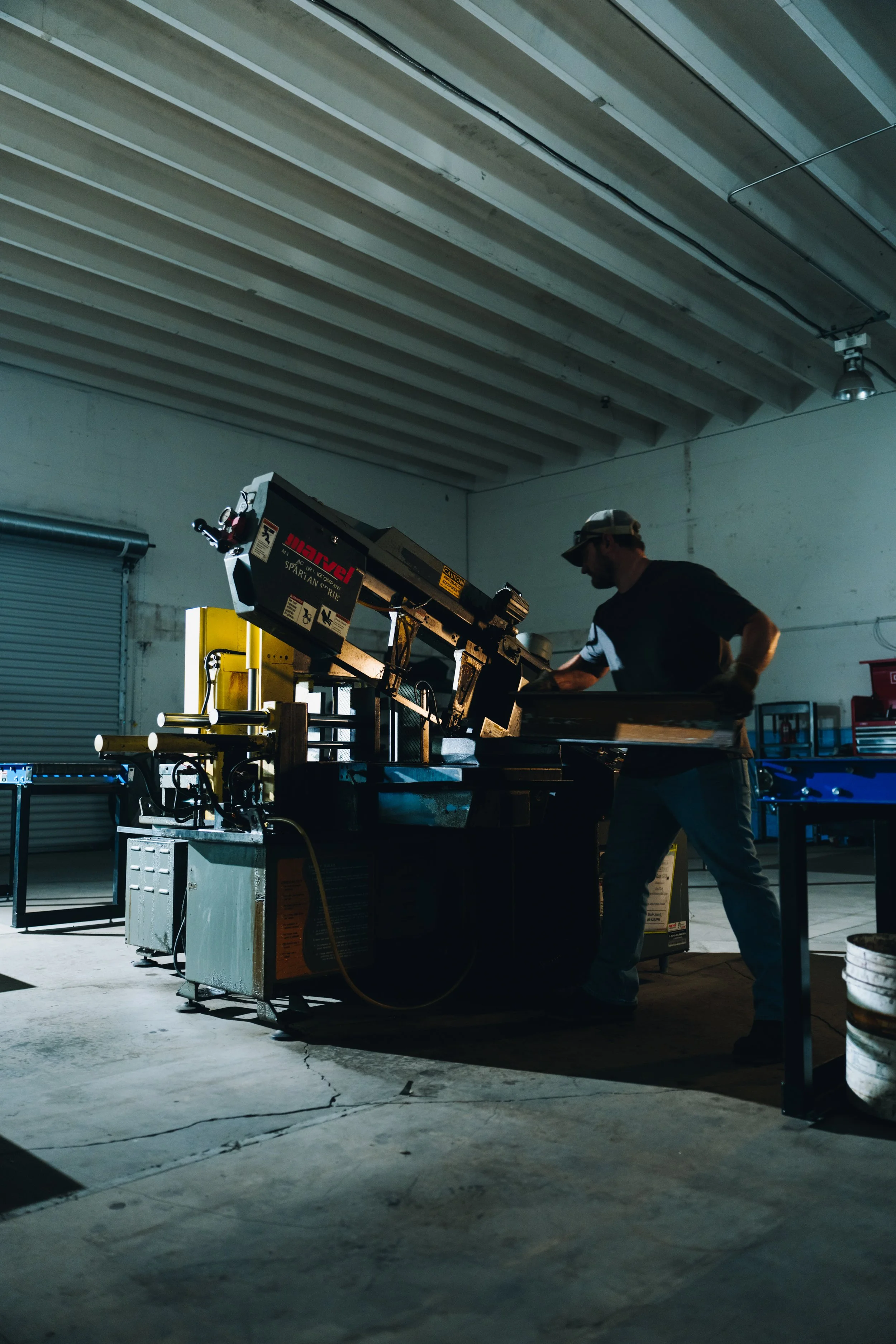 A man is working on a large metal machine in an industrial workshop with a concrete floor and exposed ceiling beams.