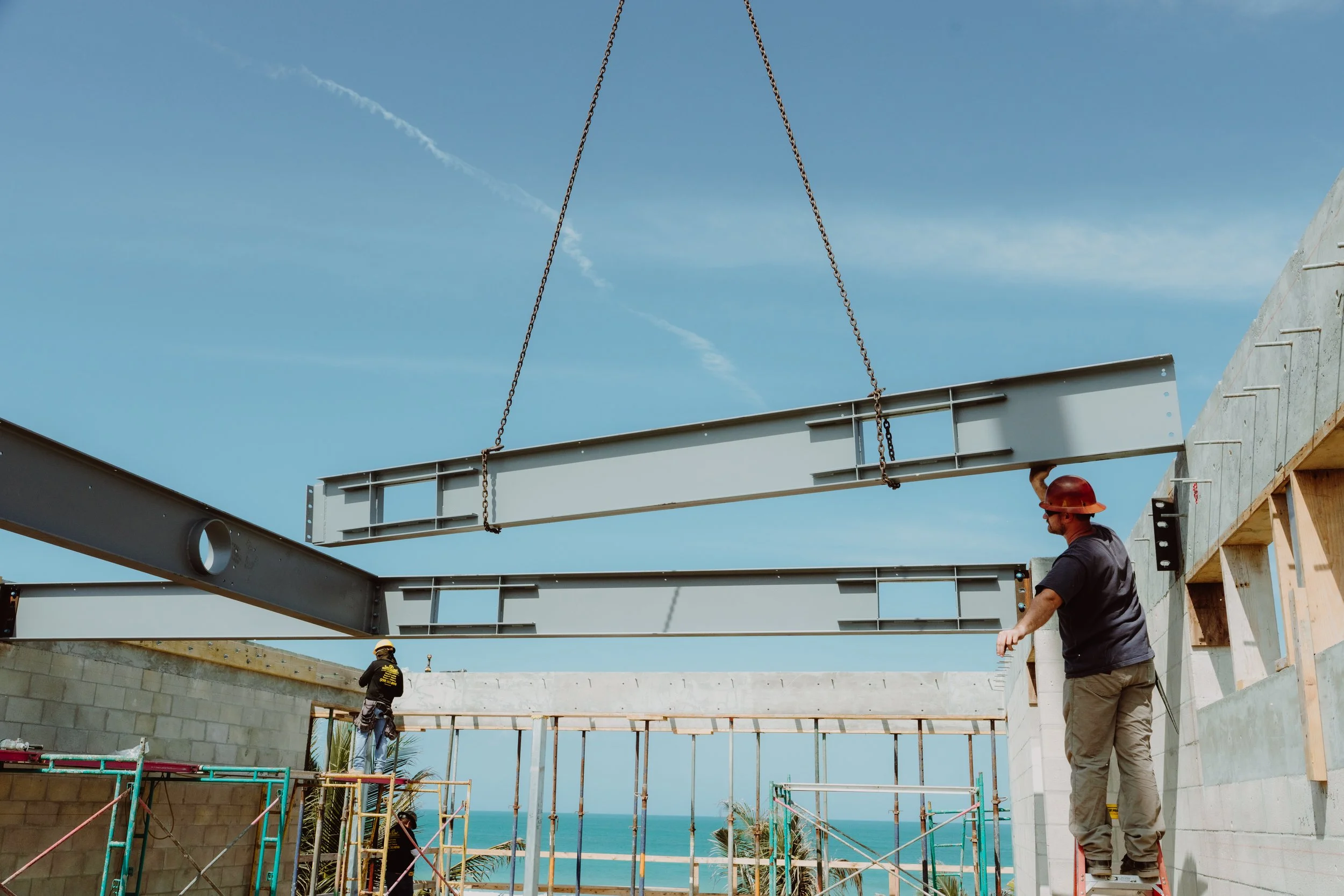 Construction workers assembling a steel beam on a building under construction near the ocean, with a clear blue sky in the background.