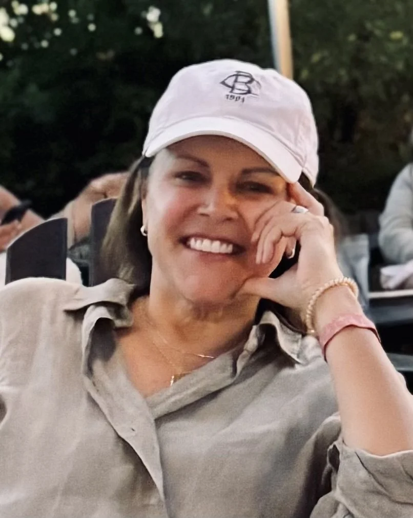 A woman smiling and wearing a light-colored shirt and a white baseball cap, sitting outdoors in a social setting.