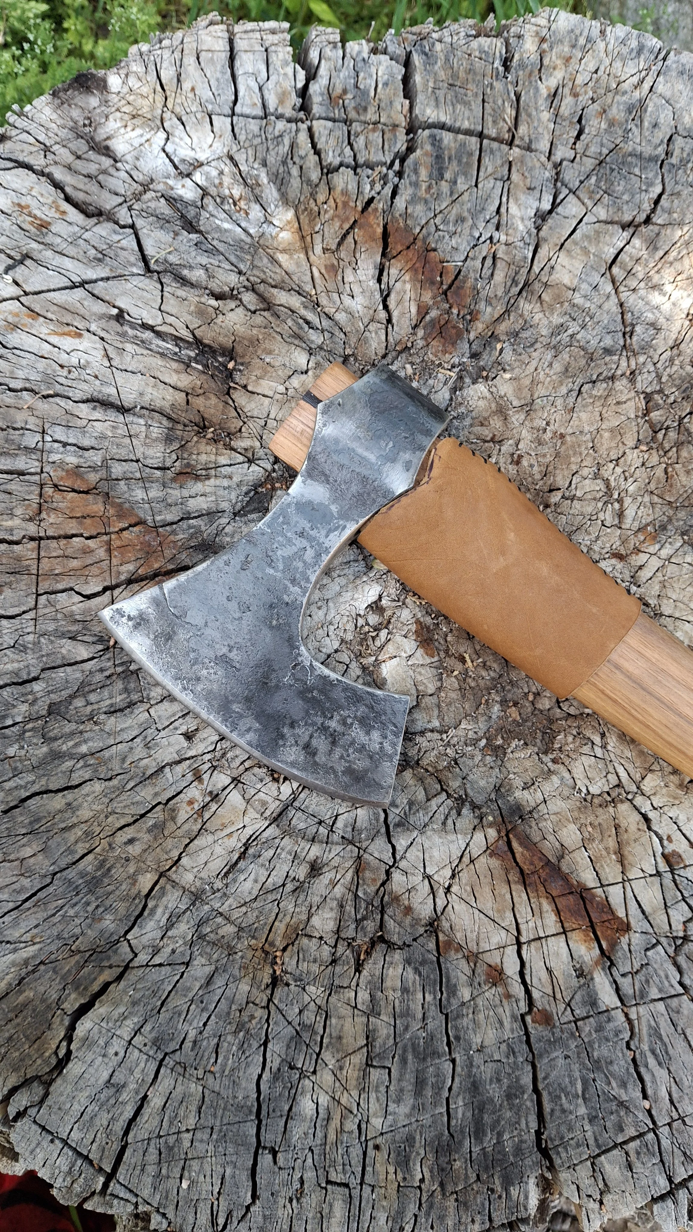 A viking style bearded axe with a wooden handle resting on a weathered, cracked tree stump.