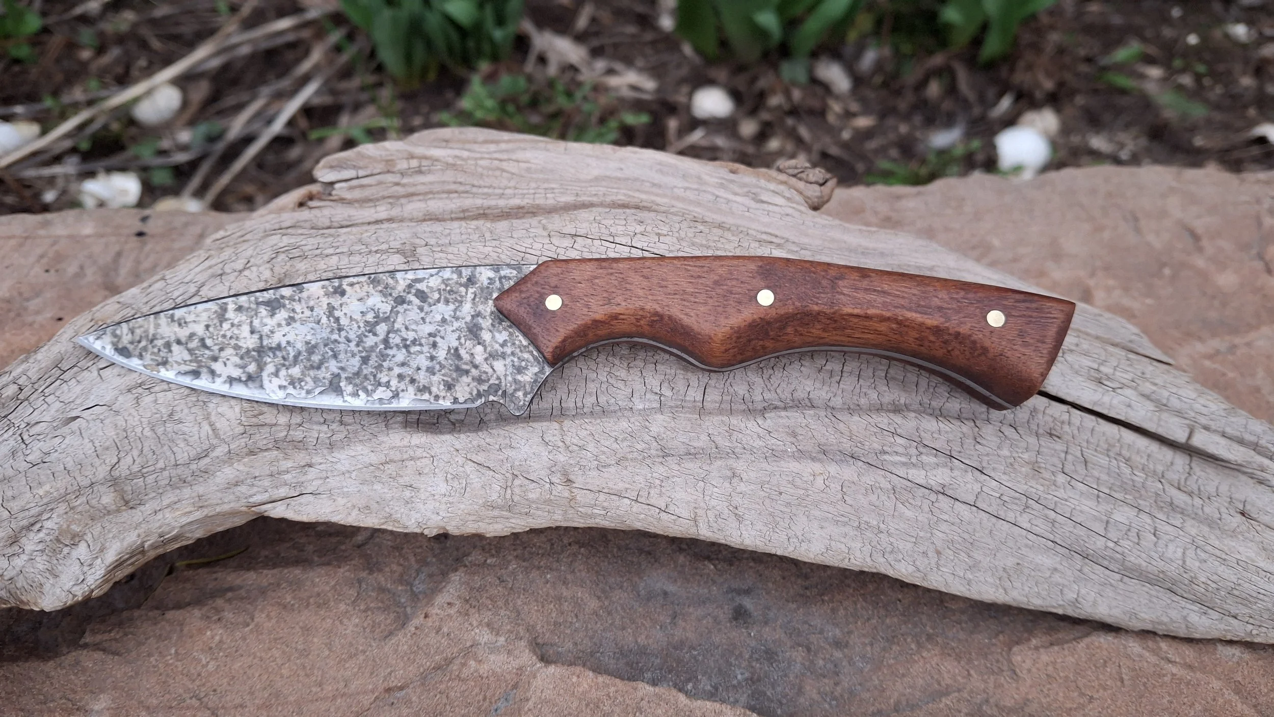 A knife with a textured metal blade and a wooden handle resting on a weathered, cracked piece of driftwood outdoors.