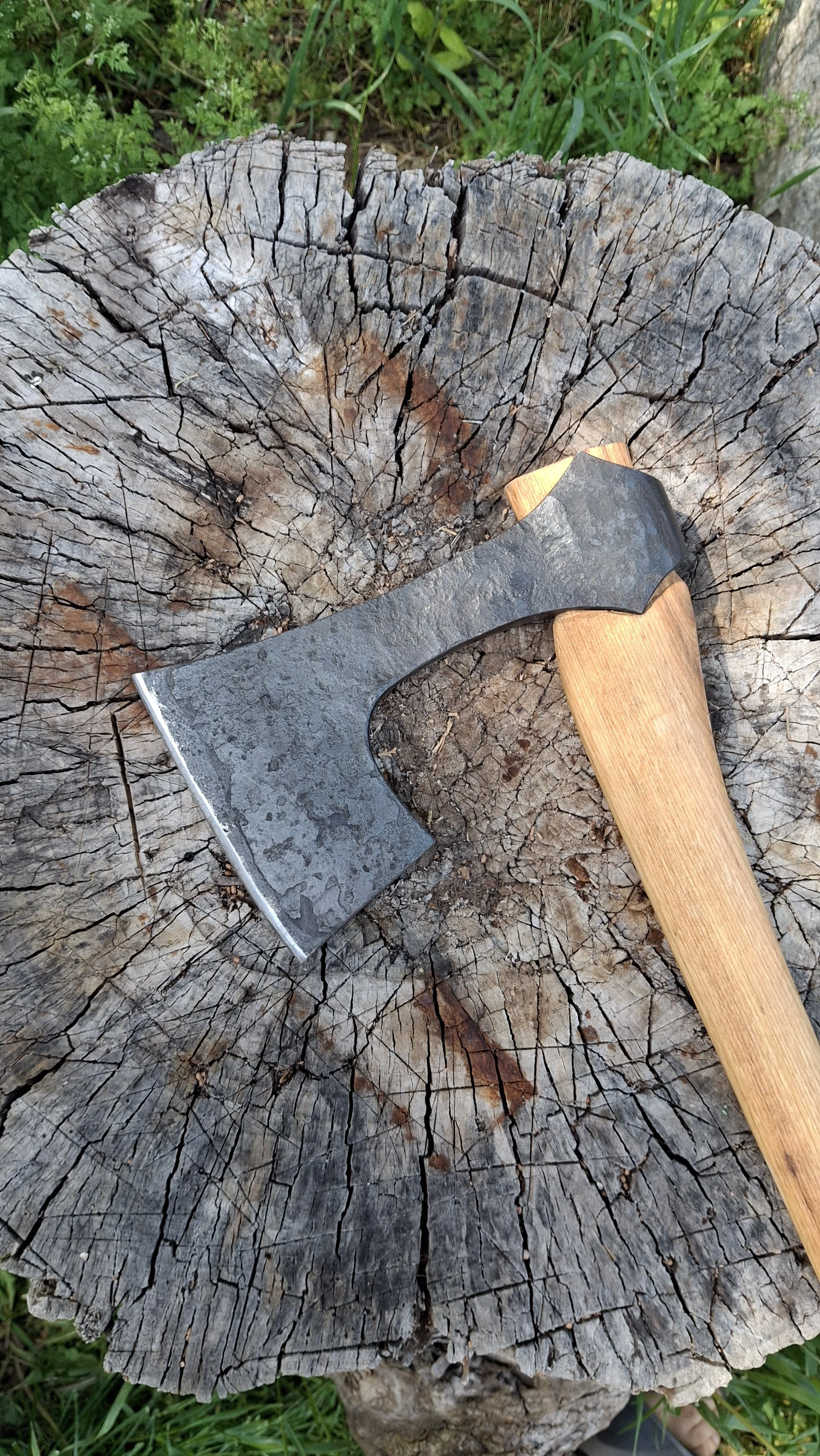 A viking style bearded axe with a wooden handle resting on the surface of a weathered tree stump with deep cracks.