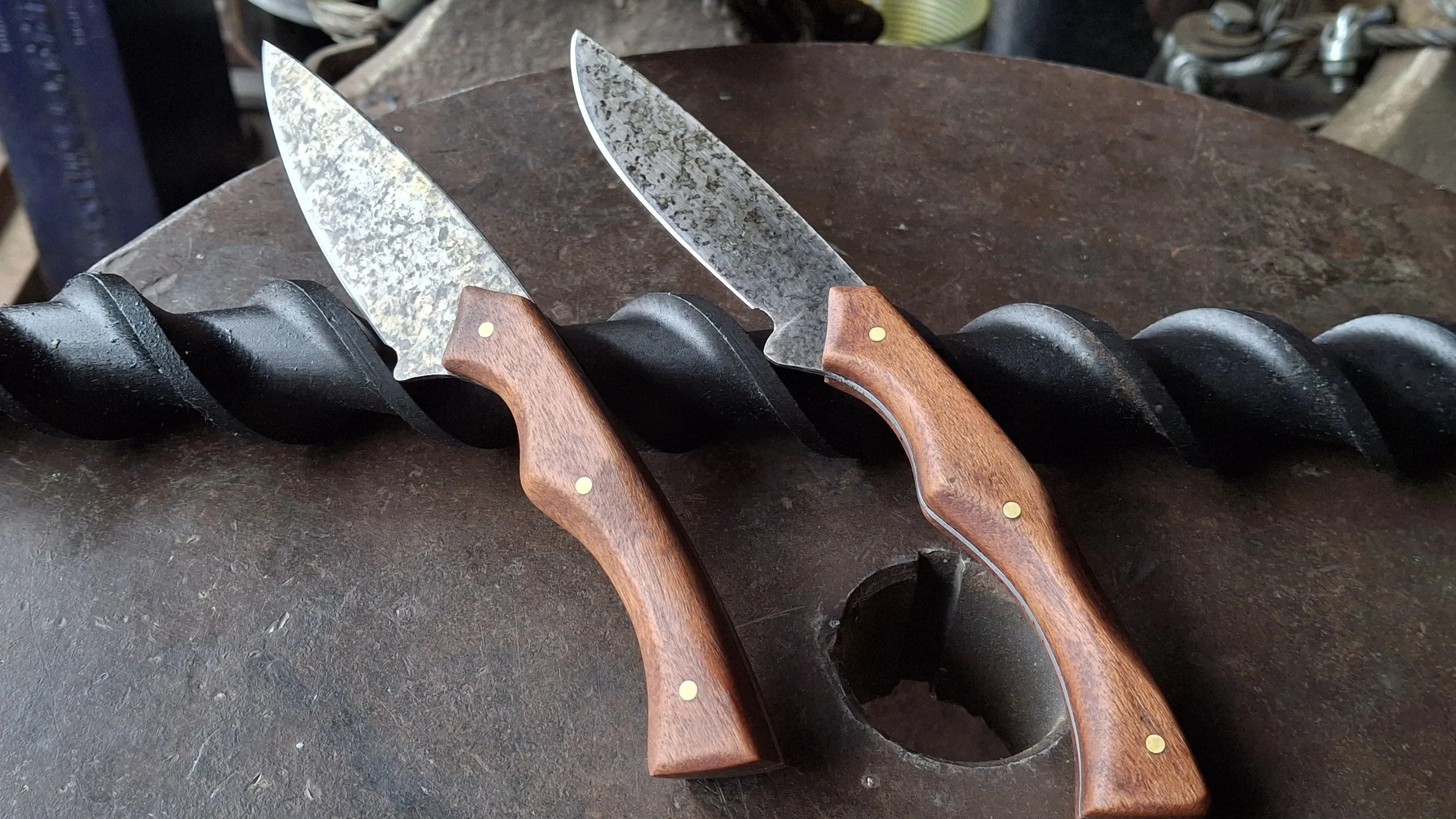 Three kitchen knives with wooden handles resting on a black metal drill just outside, with a blurry background of garden and tools.