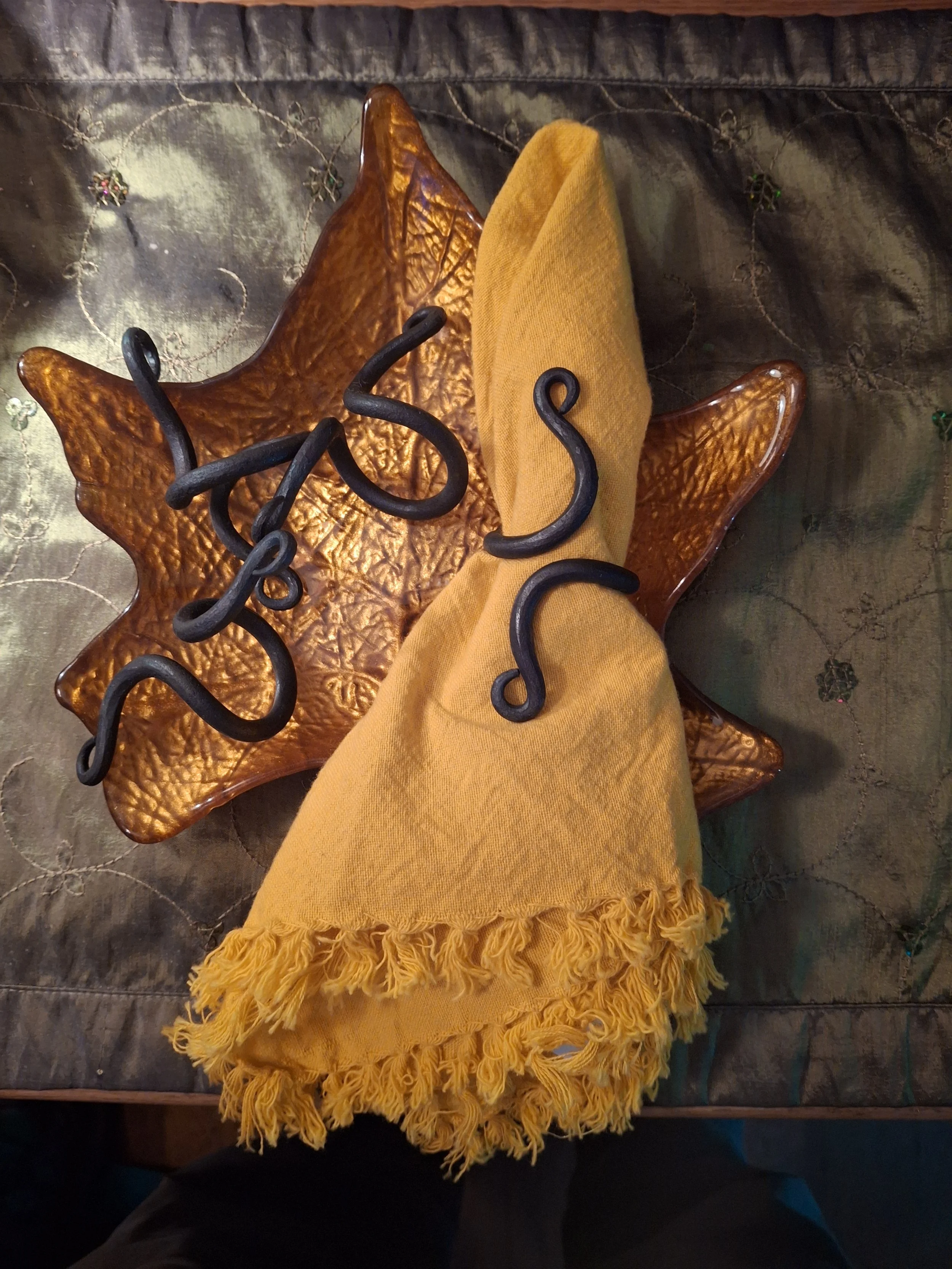 A glass bowl with a textured, star-shaped design, holding a folded yellow cloth and a black wrought iron decoration with curly details, on a dark embroidered fabric surface.