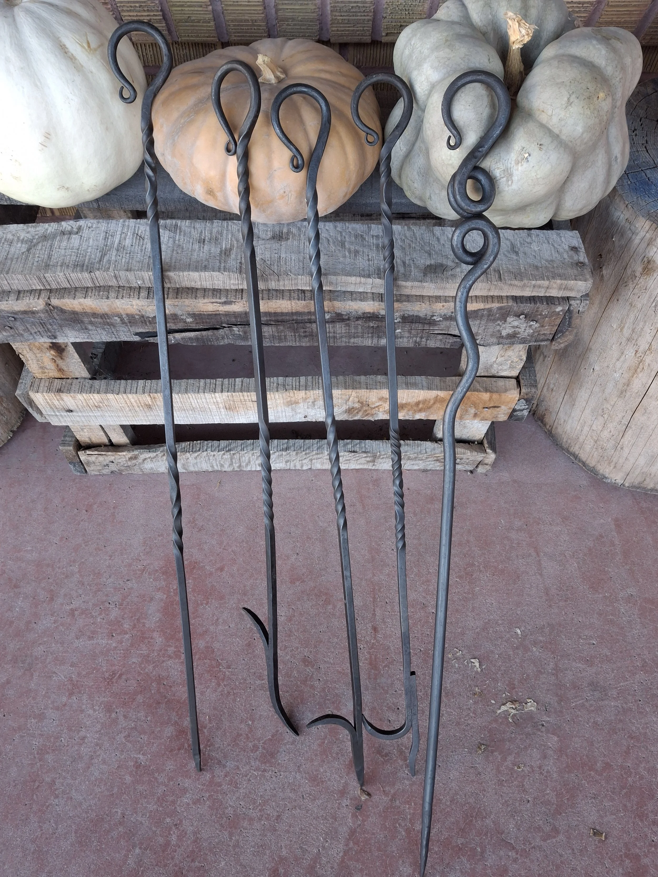 Four metal fireplace tools with twisted and curly design, resting against a wooden pallet with pumpkins and a wooden surface in the background.