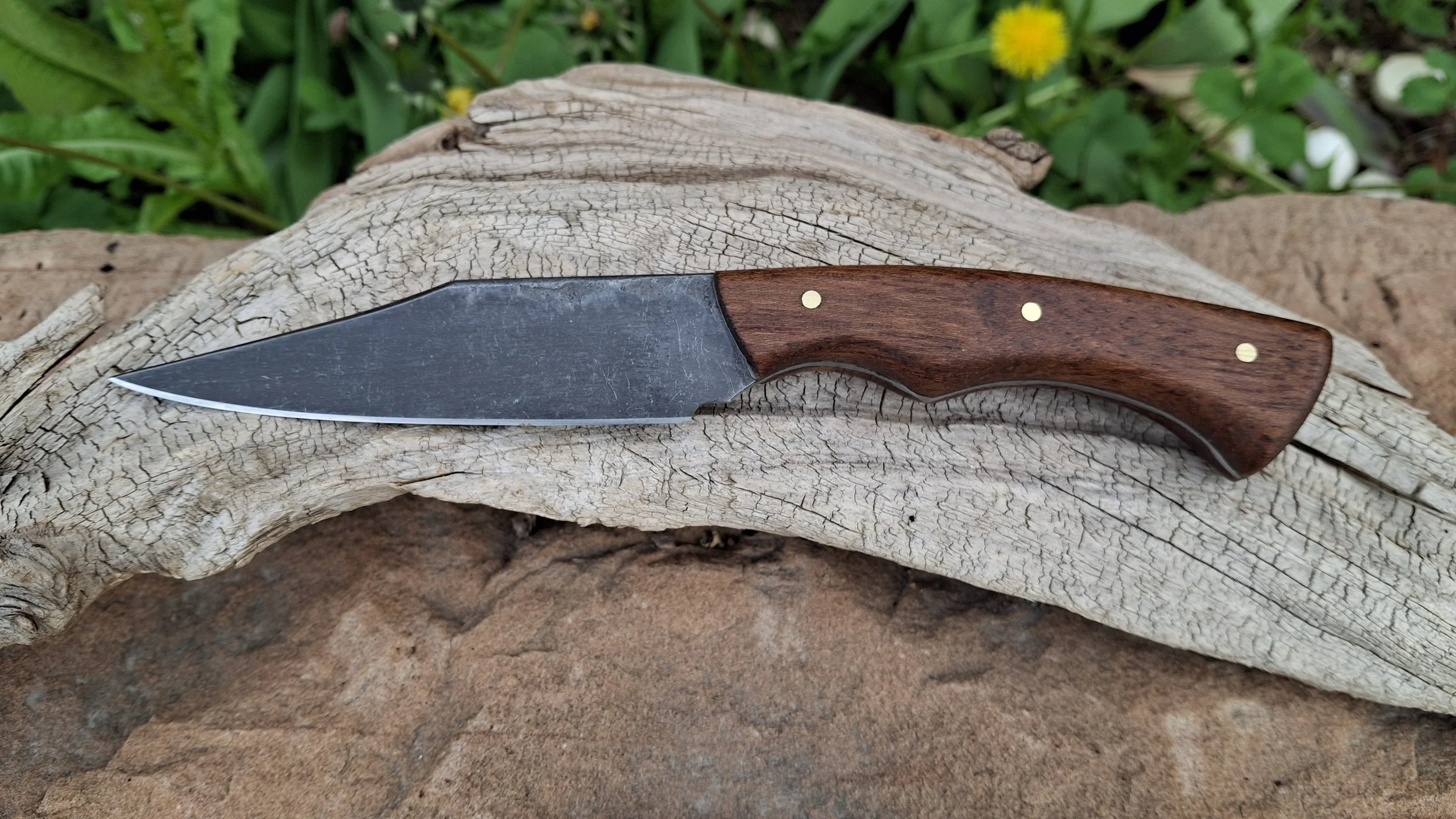 A fixed-blade knife with a wooden handle and a slightly curved steel blade, resting on a piece of weathered wood outdoors with greenery and yellow flowers in the background.