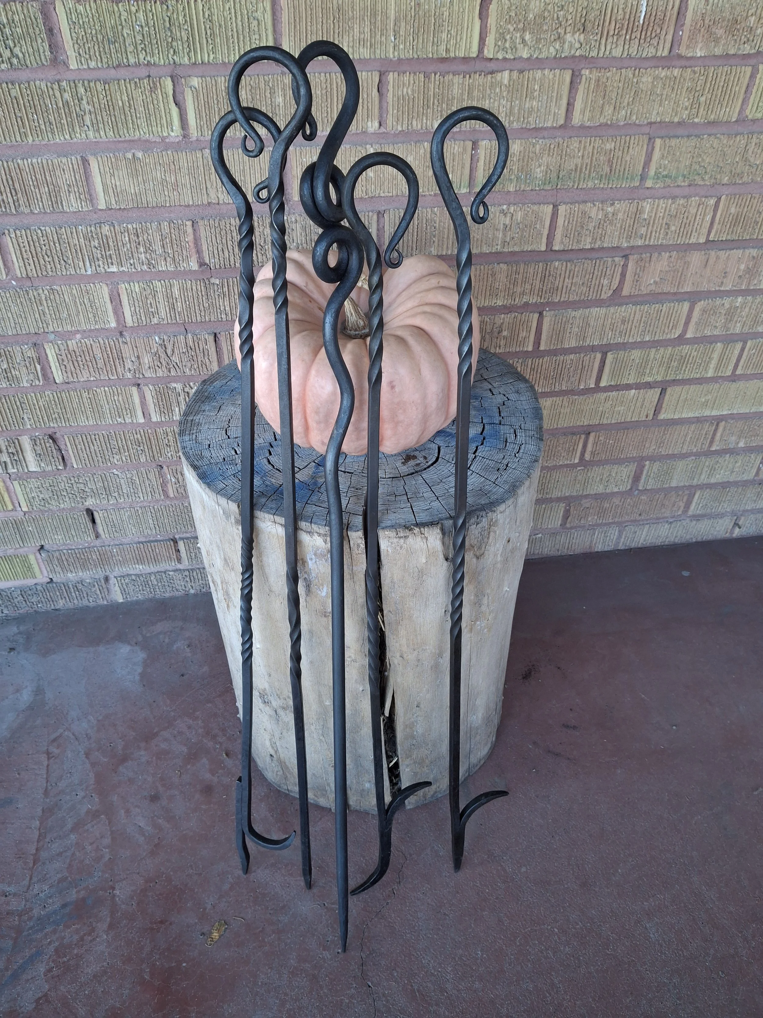A large, pink pumpkin on a wooden stump with four black wrought iron fireplace tools leaning against it, set against a brick wall.