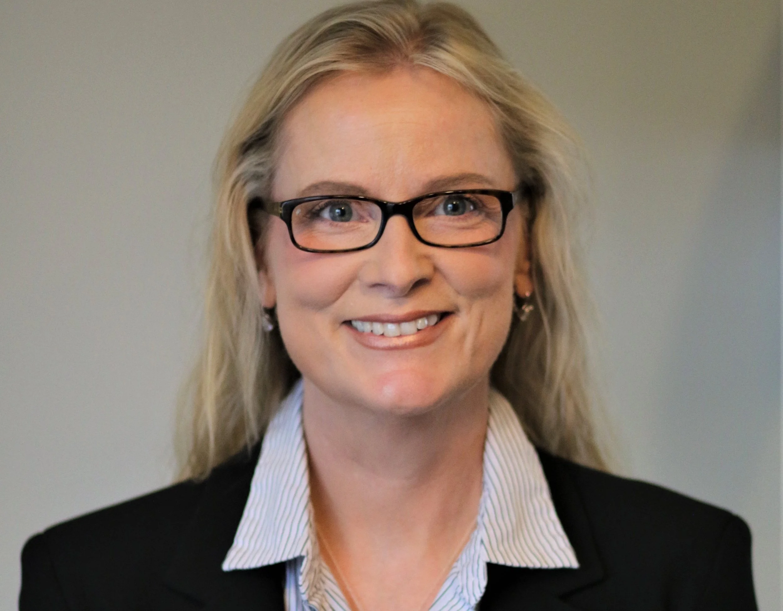Close-up of a smiling woman with blonde hair, wearing black glasses, a white collared shirt with thin blue stripes, and a black blazer against a plain background.