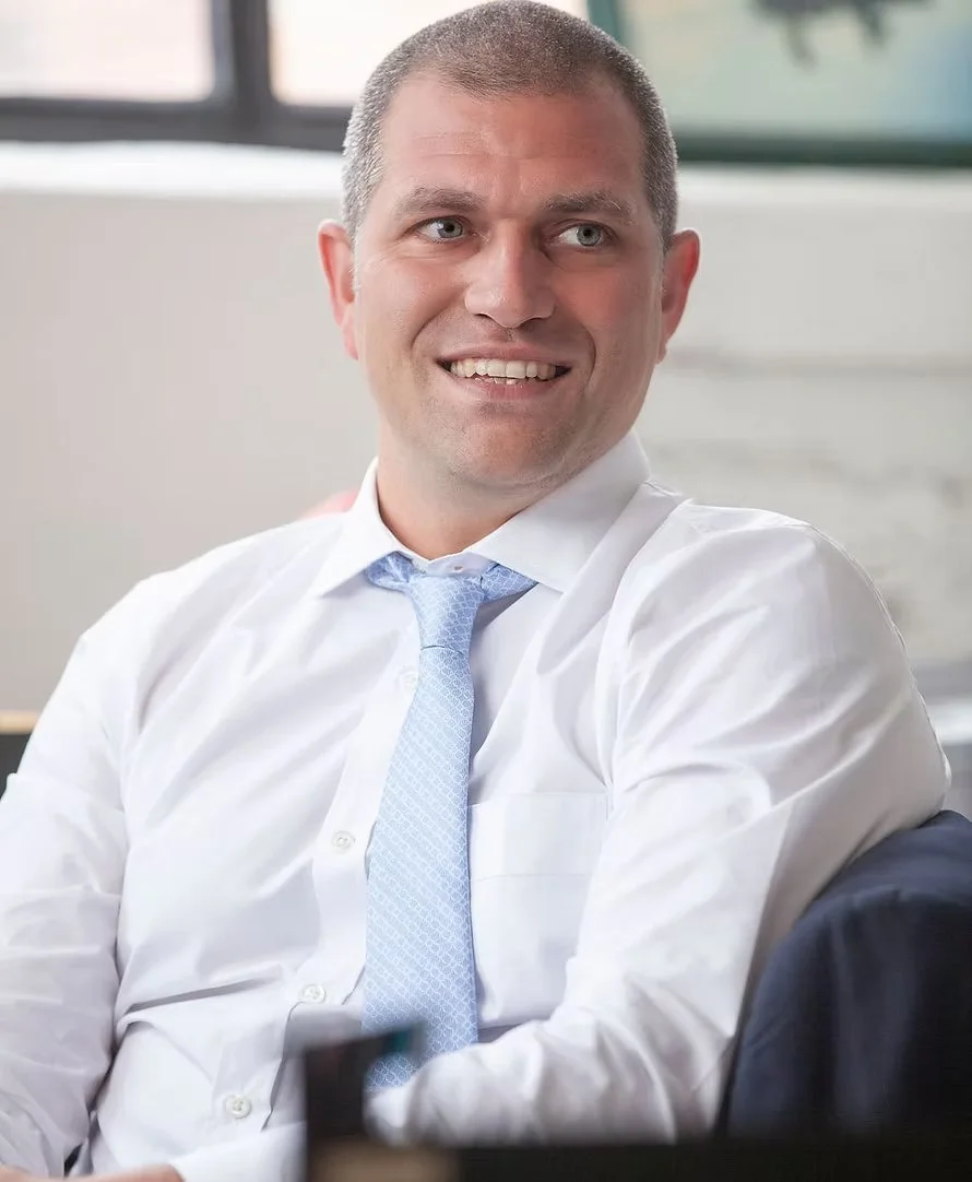 A man with short hair wearing a white dress shirt and a light blue tie, smiling while seated indoors.