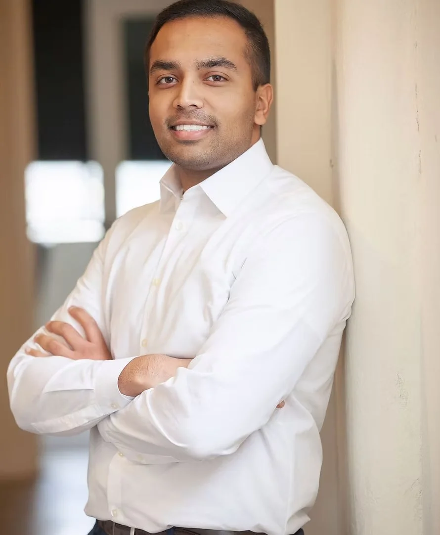 A man with short dark hair, wearing a white button-down shirt, standing with arms crossed and smiling at the camera, indoors with blurred background.