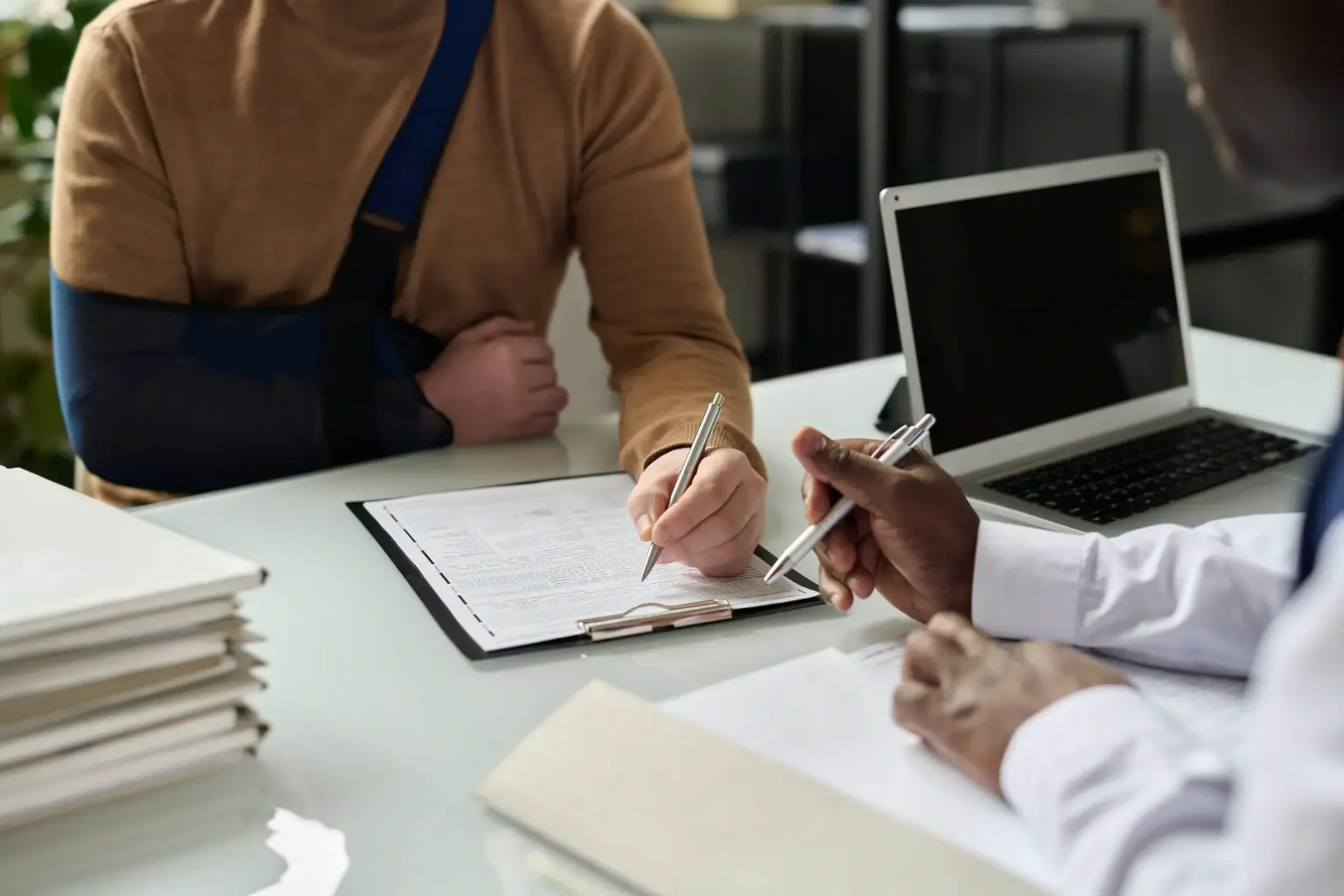 A person with a broken arm in a cast sitting at a table, signing a document with another person, who is holding a pen. There are stacks of papers, a laptop, and a clipboard on the table.