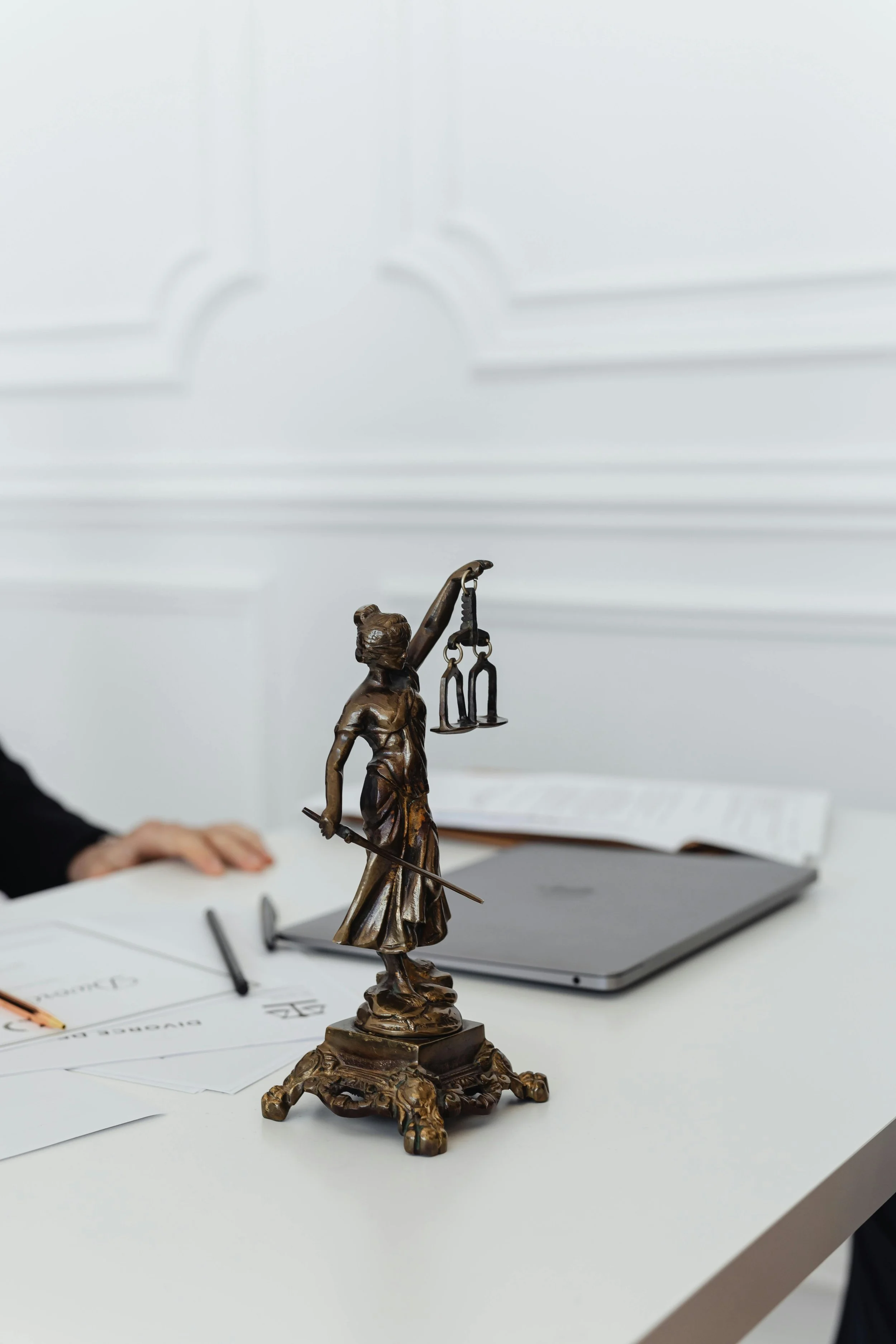 A bronze statue of Lady Justice holding a scale in her left hand and a sword in her right hand, placed on a white table with documents, a pen, a pair of glasses, a closed laptop, and an open book in the background.