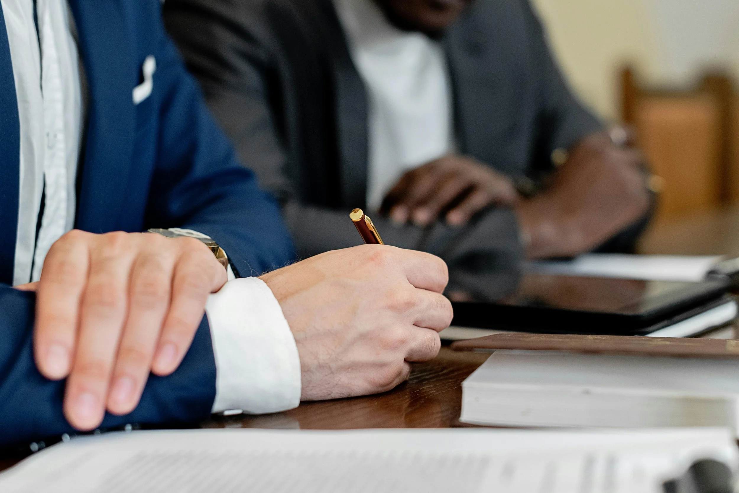 Close-up of two people sitting at a table in a meeting, one taking notes with a pen, and the other with crossed arms, with notebooks and documents on the table.