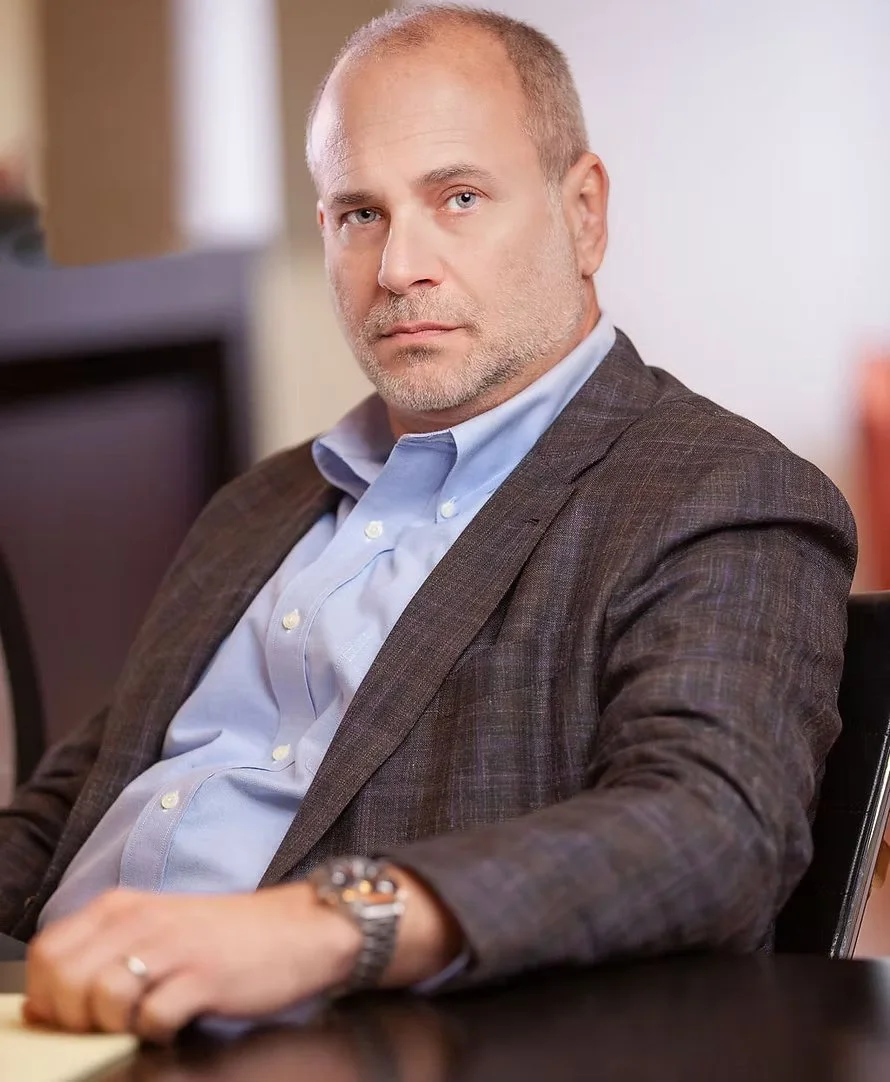 A middle-aged man with a serious expression, wearing a blue dress shirt and a brown blazer, sitting at a desk in an office environment.