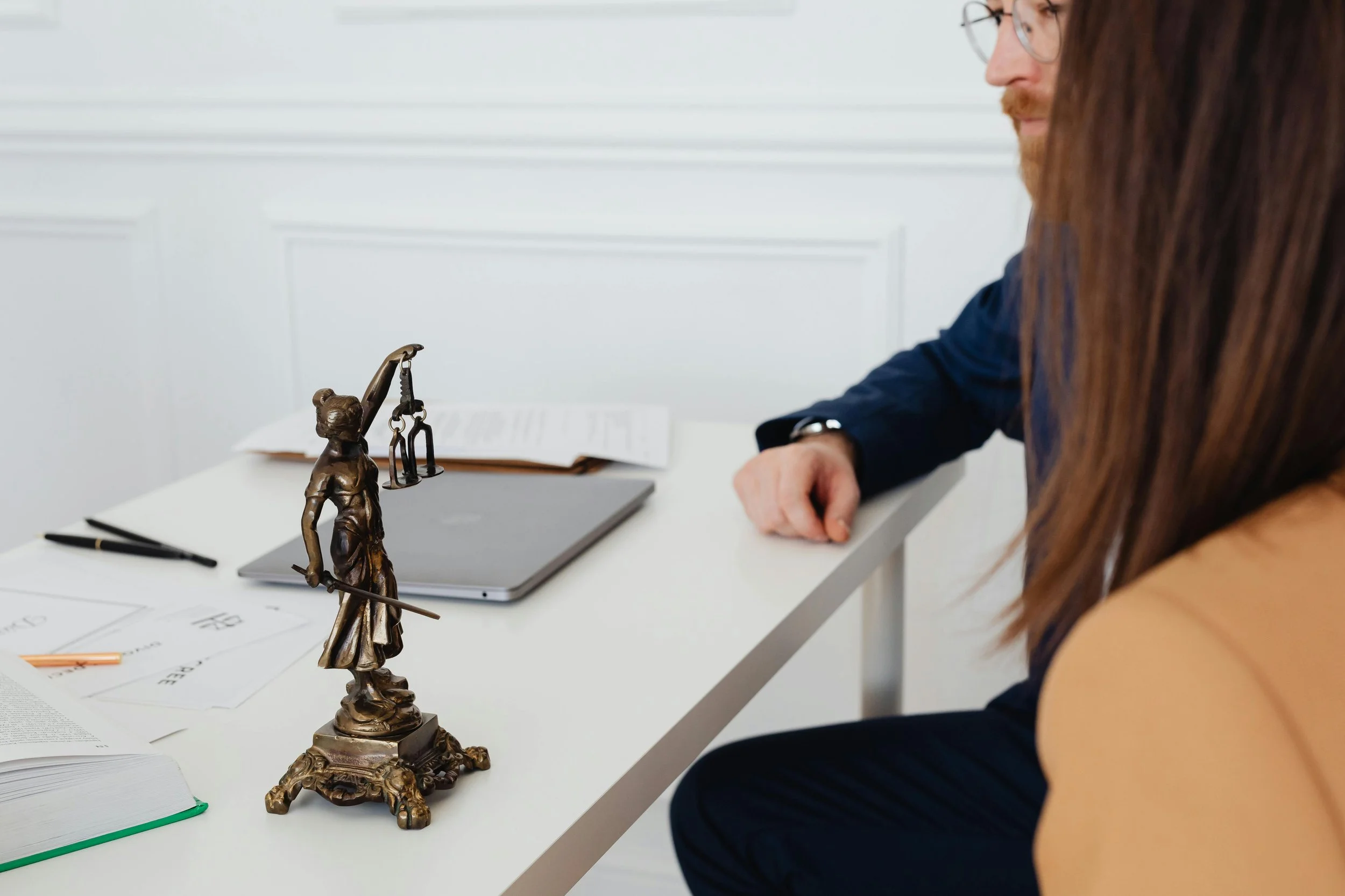 Close-up of a bronze statue of Lady Justice on a white desk, with a person sitting nearby.