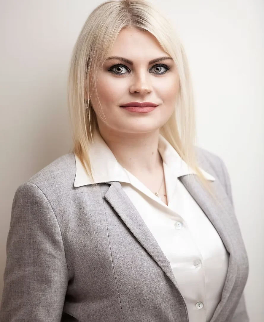 Portrait of a blonde woman in a gray blazer and white shirt standing against a plain light background.