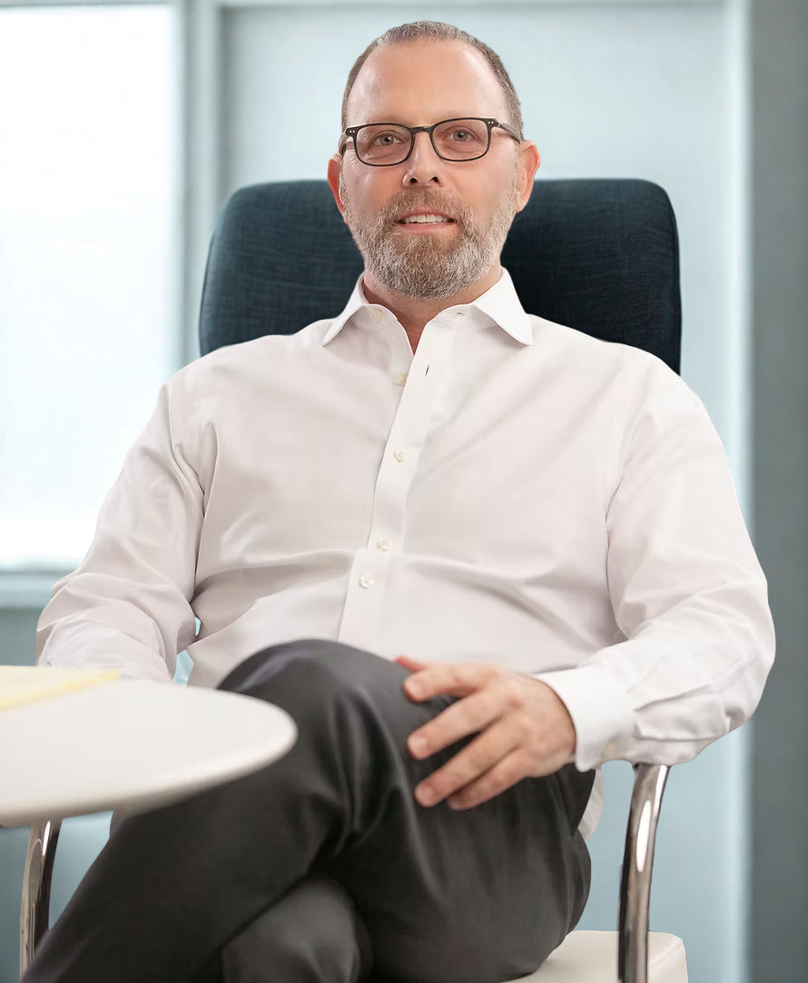 A man with glasses and a beard, wearing a white dress shirt, sitting in an office.