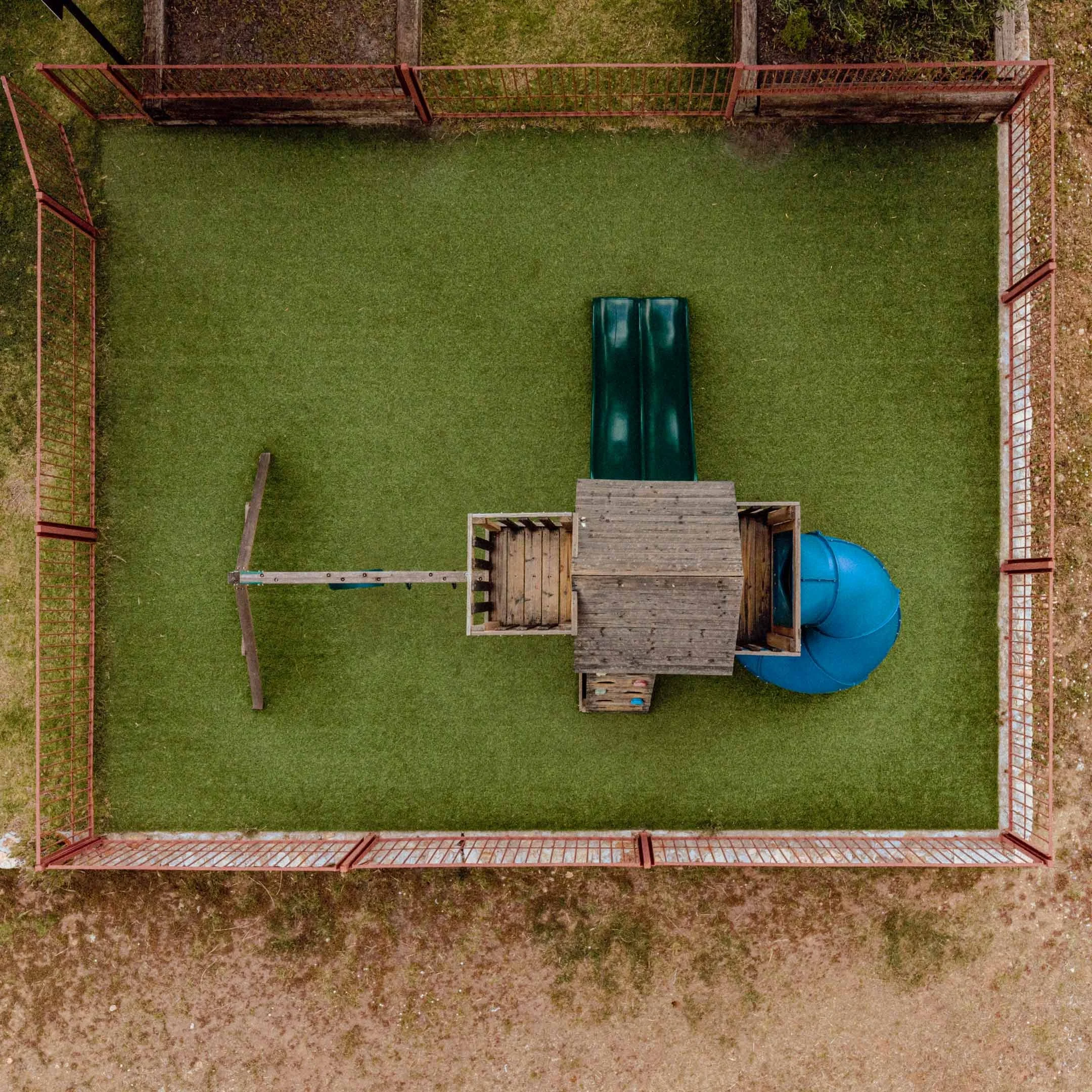 Vista aérea de un área de juego infantil con un tobogán azul y una estructura de madera rodeada por una cerca de metal roja.