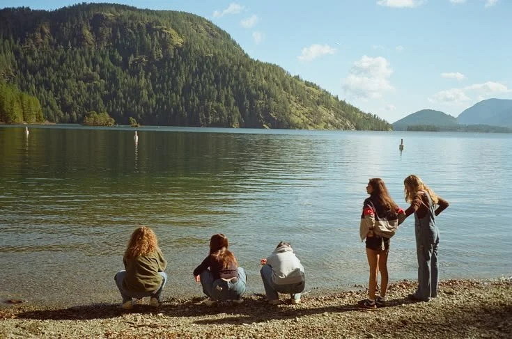 Five people sitting and standing on a rocky shore by a lake, with forested hills in the background.