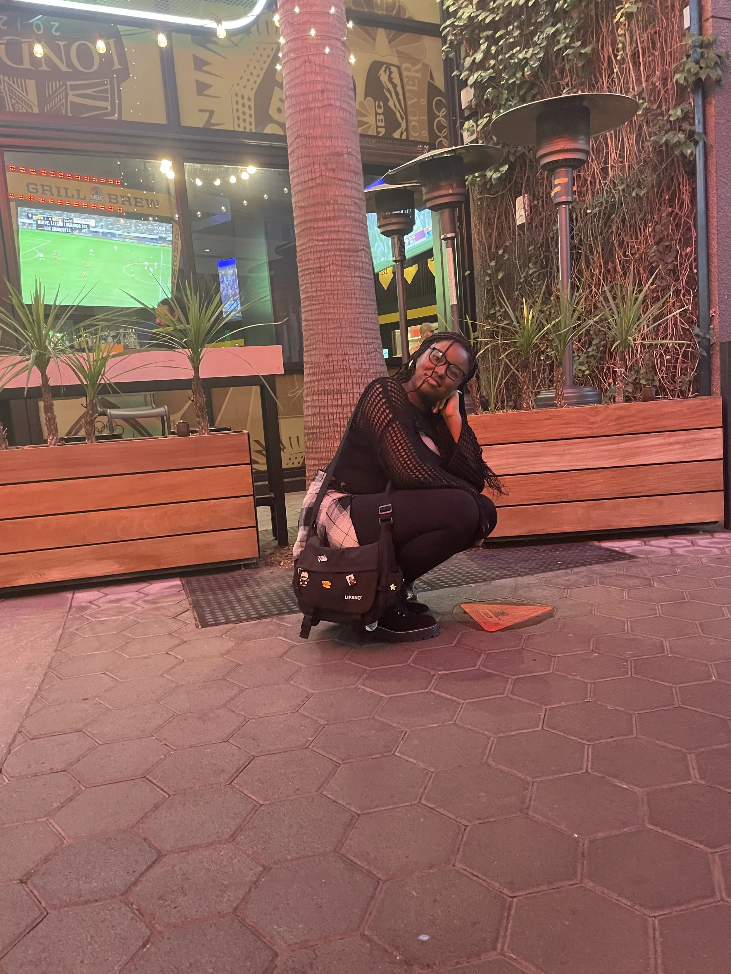 A woman crouching on a sidewalk outside a restaurant or bar, with a large glass window showing a television screen playing a sports game, potted plants, and outdoor heaters in the background. She is wearing black clothing, glasses, and sneakers, with a backpack beside her.