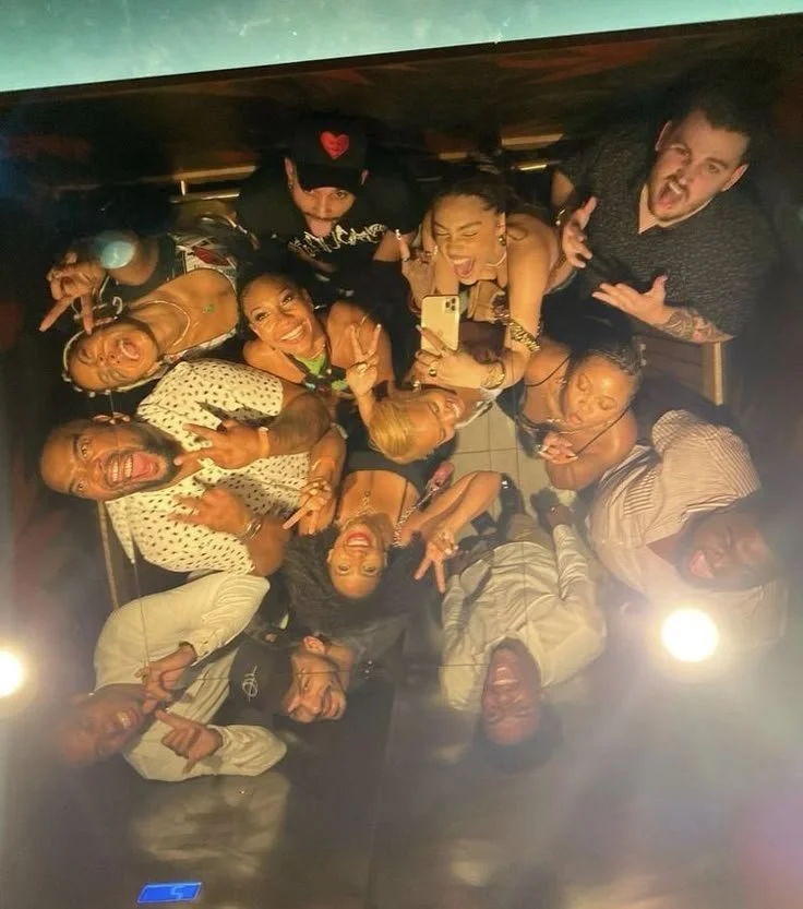 A group of diverse people are taking a selfie from an overhead perspective, all smiling and making expressive gestures, in a cozy indoor setting.