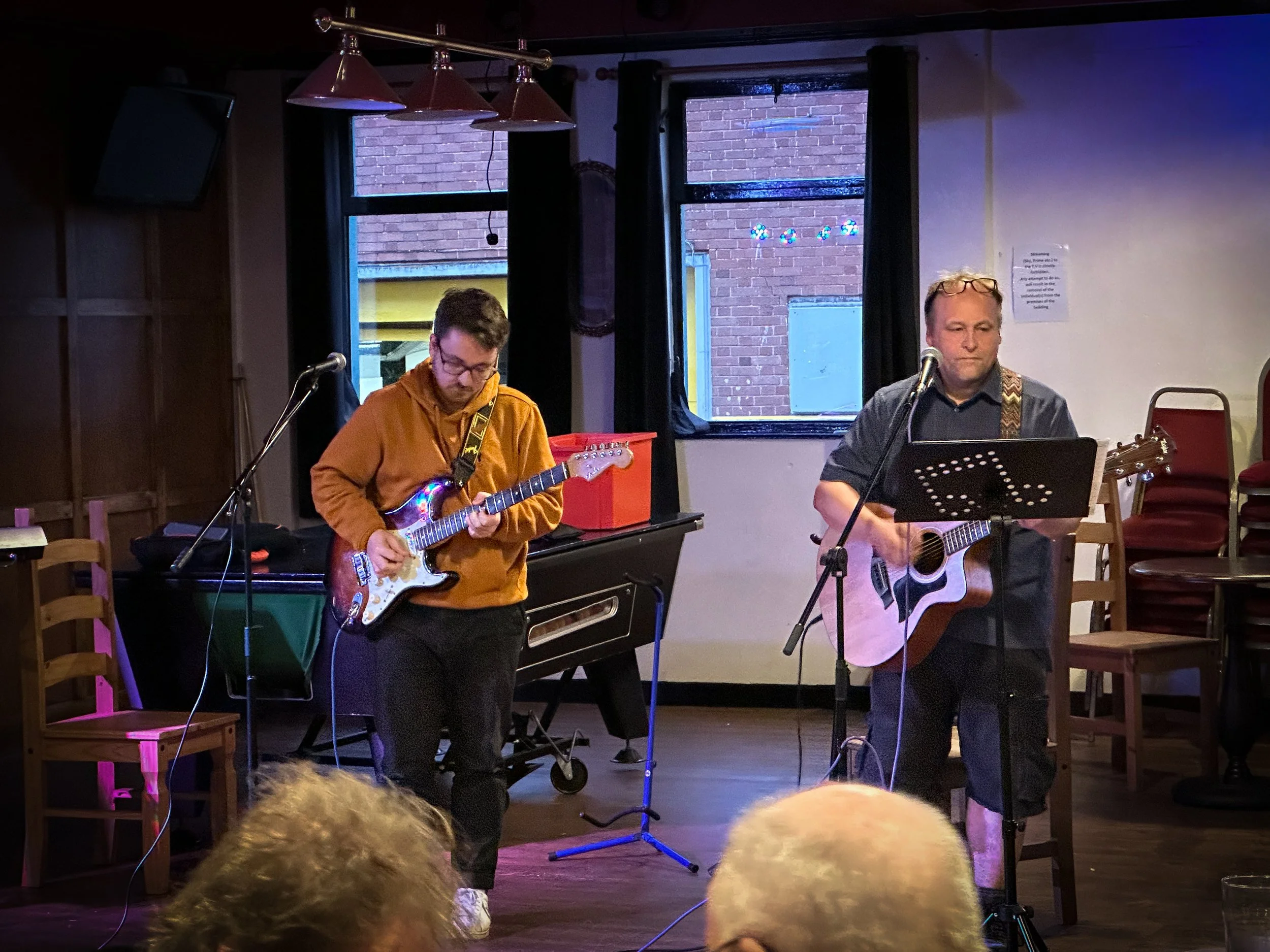 Two musicians perform on stage with guitars, one wearing glasses and a dark shirt, and the other in an orange hoodie, in a dimly lit room with a black curtain, a window, and a wall-mounted TV.