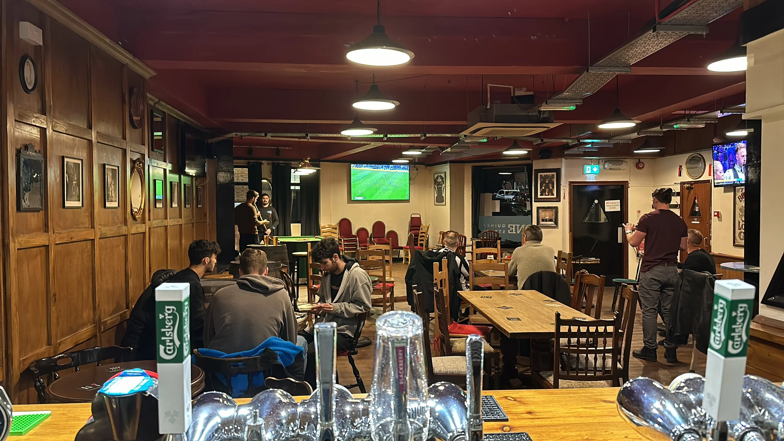 People watching a soccer game on multiple television screens inside a cozy pub with wood-paneled walls and framed pictures, some sitting at tables, others standing and socializing.