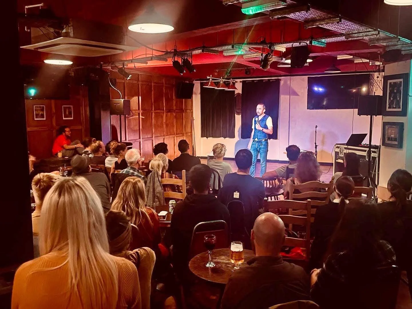 A comedy show taking place on a small stage in a cozy, dimly lit bar or club. A man is performing on stage, holding a microphone, with an audience seated at tables watching him. Some guests have drinks, and the background features black curtains and 