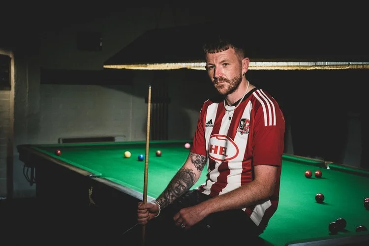 A man sitting at a pool table holding a cue stick, wearing a red and white striped soccer jersey, with tattoos on his arm and a serious expression.