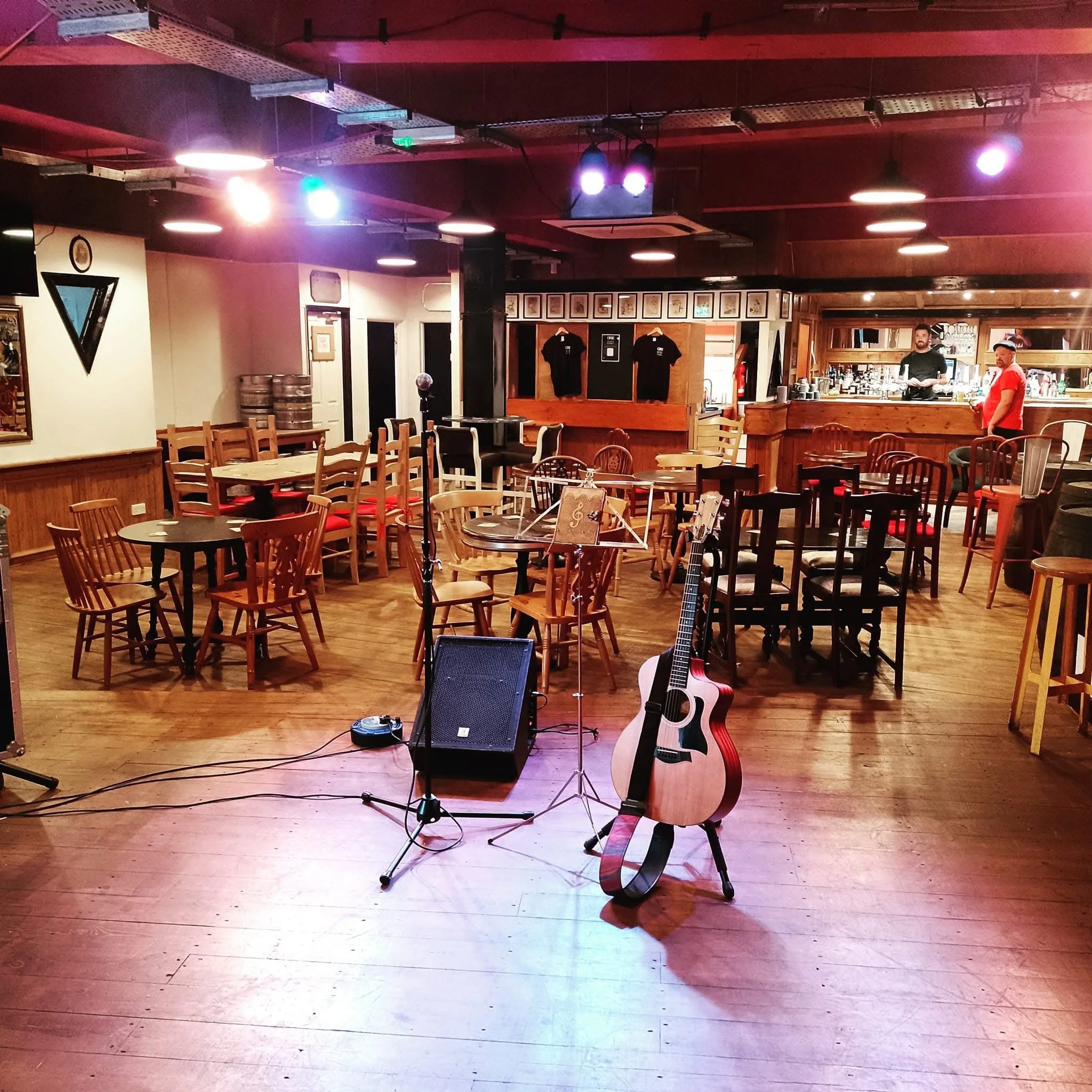 Empty stage with musical equipment including guitar, amplifier, and microphone, in front of a bar area with two staff members behind the counter. Several chairs and tables are arranged in the foreground, with colorful lighting effects on the ceiling.