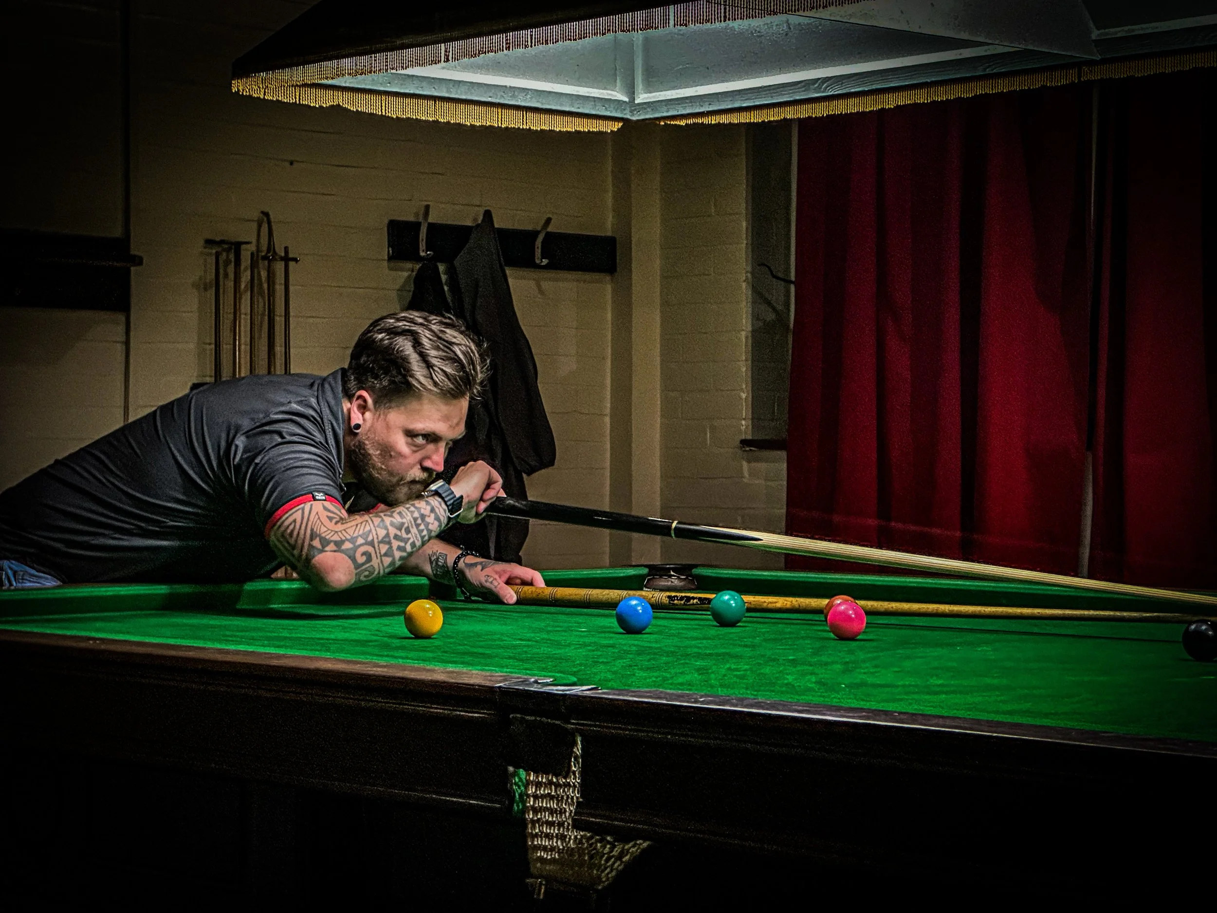 A man with tattoos and earrings leaning over a pool table, aiming to take a shot. The pool table has a green felt surface with colored balls laid out, and a cue stick is in his hands. In the background, there are red curtains, hooks with black clothi