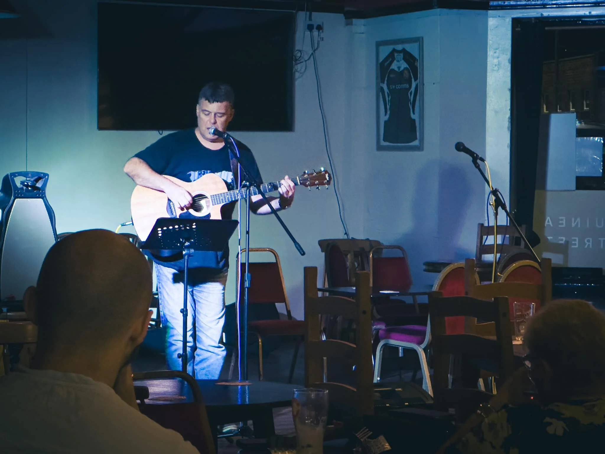 A man is playing an acoustic guitar and singing into a microphone in a dimly lit room, likely performing at a small venue or bar, with people seated at tables in the foreground.