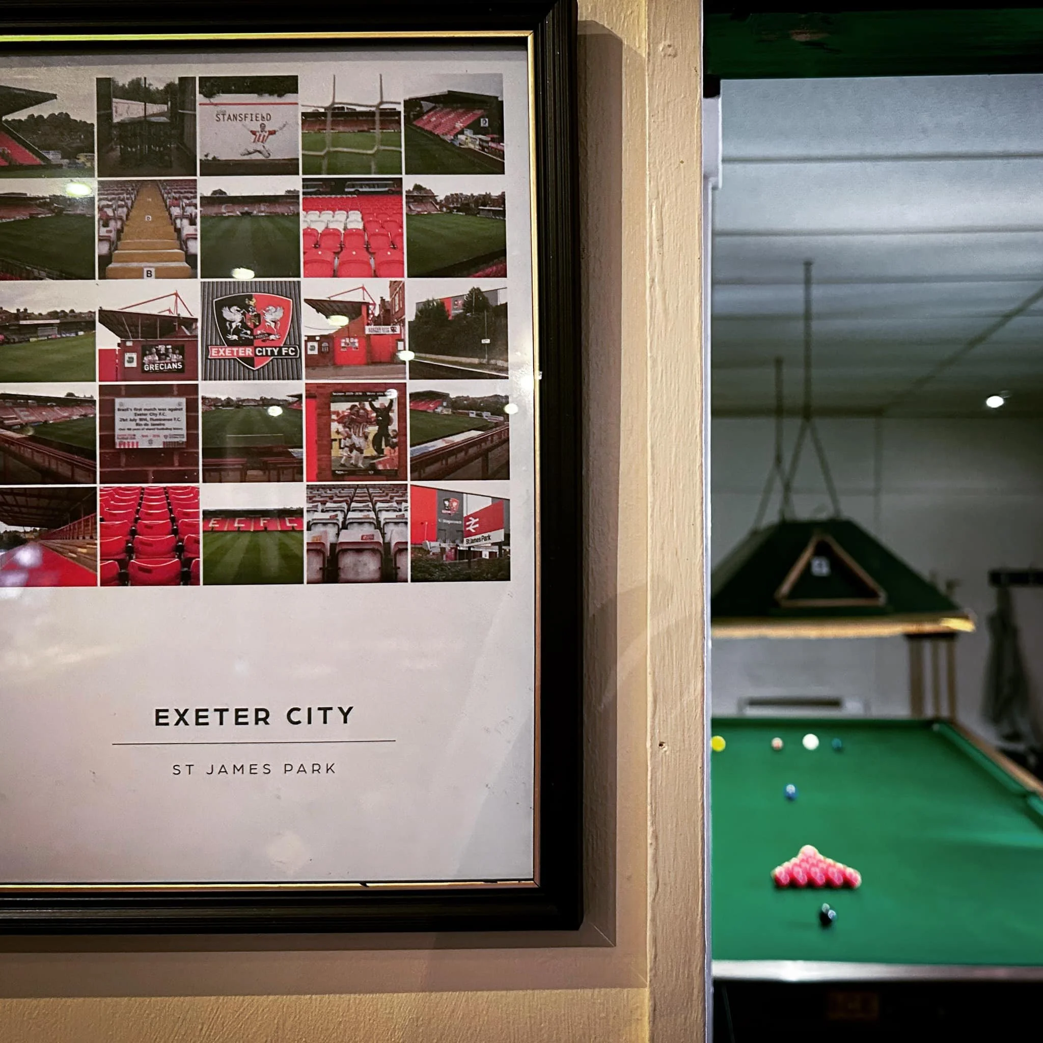 Framed collage of Exeter City football stadium and team images on a wall near a pool table with colored balls and pink chalk.