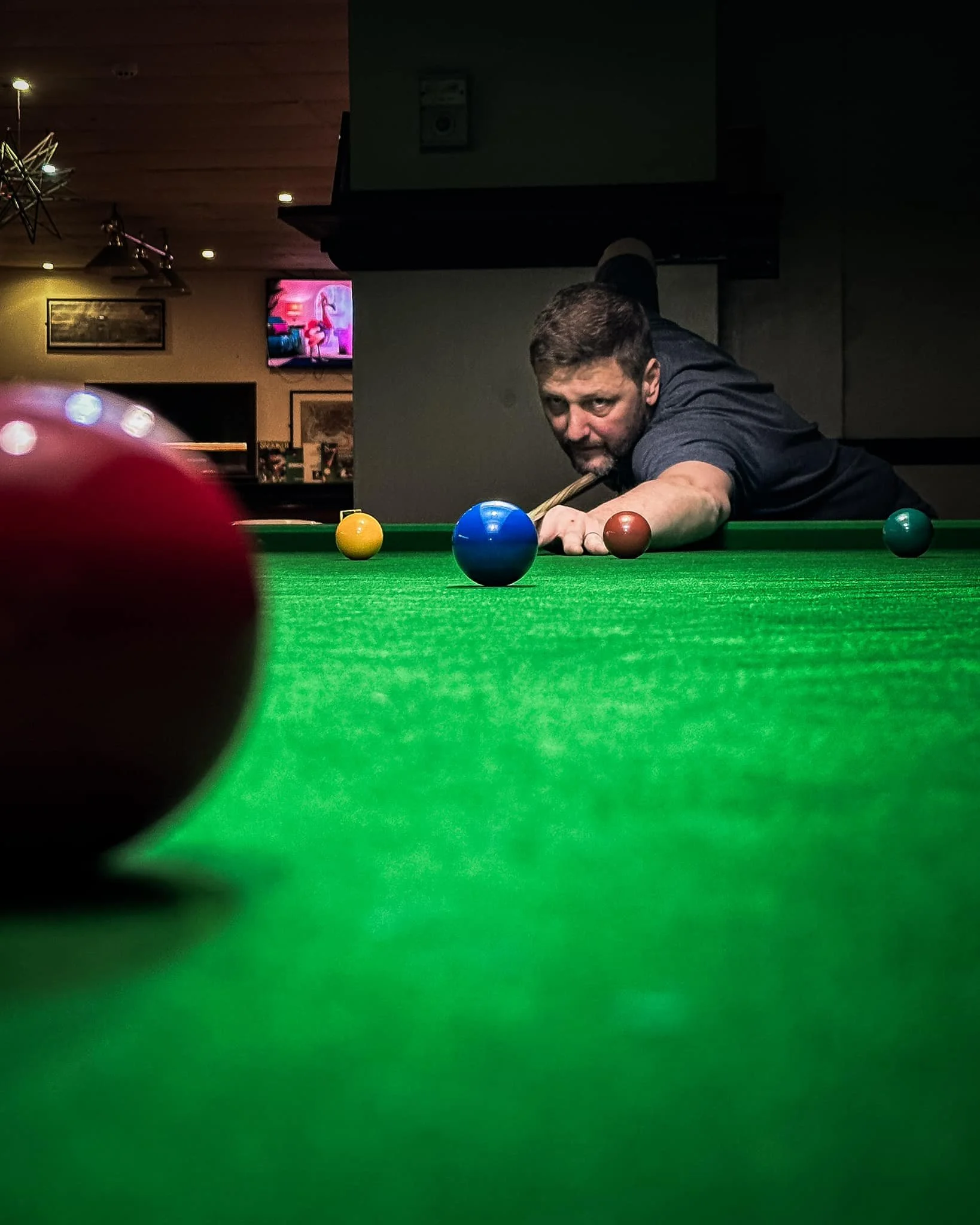 A man is playing billiards, focused on his shot, in a dimly lit pool hall with pool balls on the table and a TV showing a cartoon on the wall.