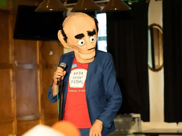 Person wearing a puppet mask of a cartoonish face, holding a microphone, standing indoors with wooden walls and black curtains.