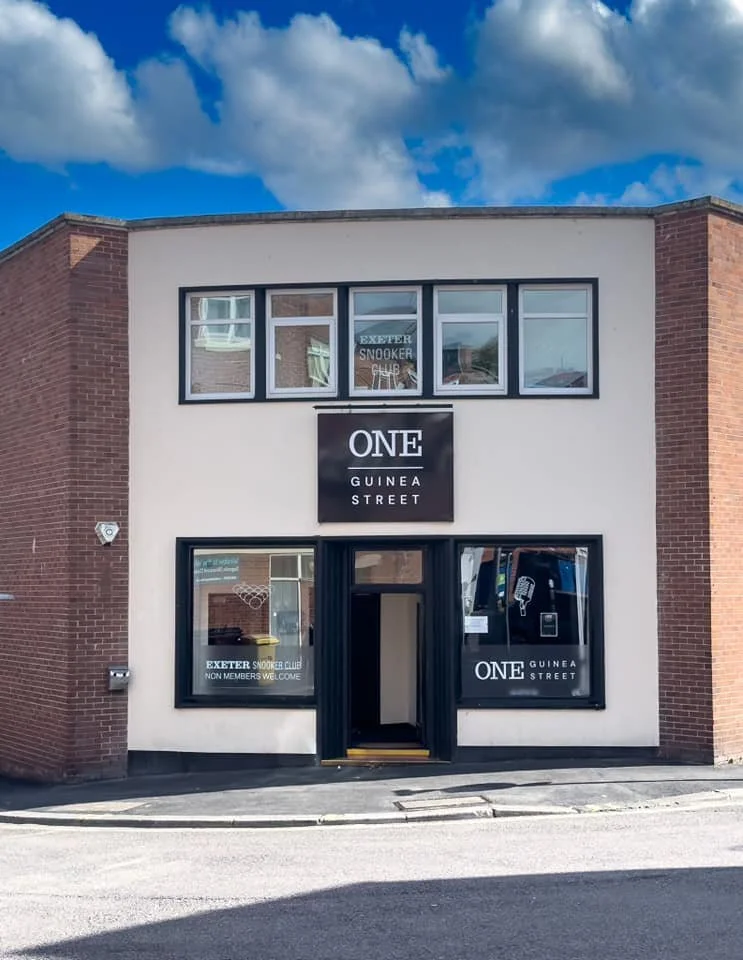 Front of a two-story building with a white facade and brick sides, featuring a sign that reads "ONE GUINEA STREET". The top window has a sign for the Exeter Snooker Club, and the lower window has a welcome message for non-members.