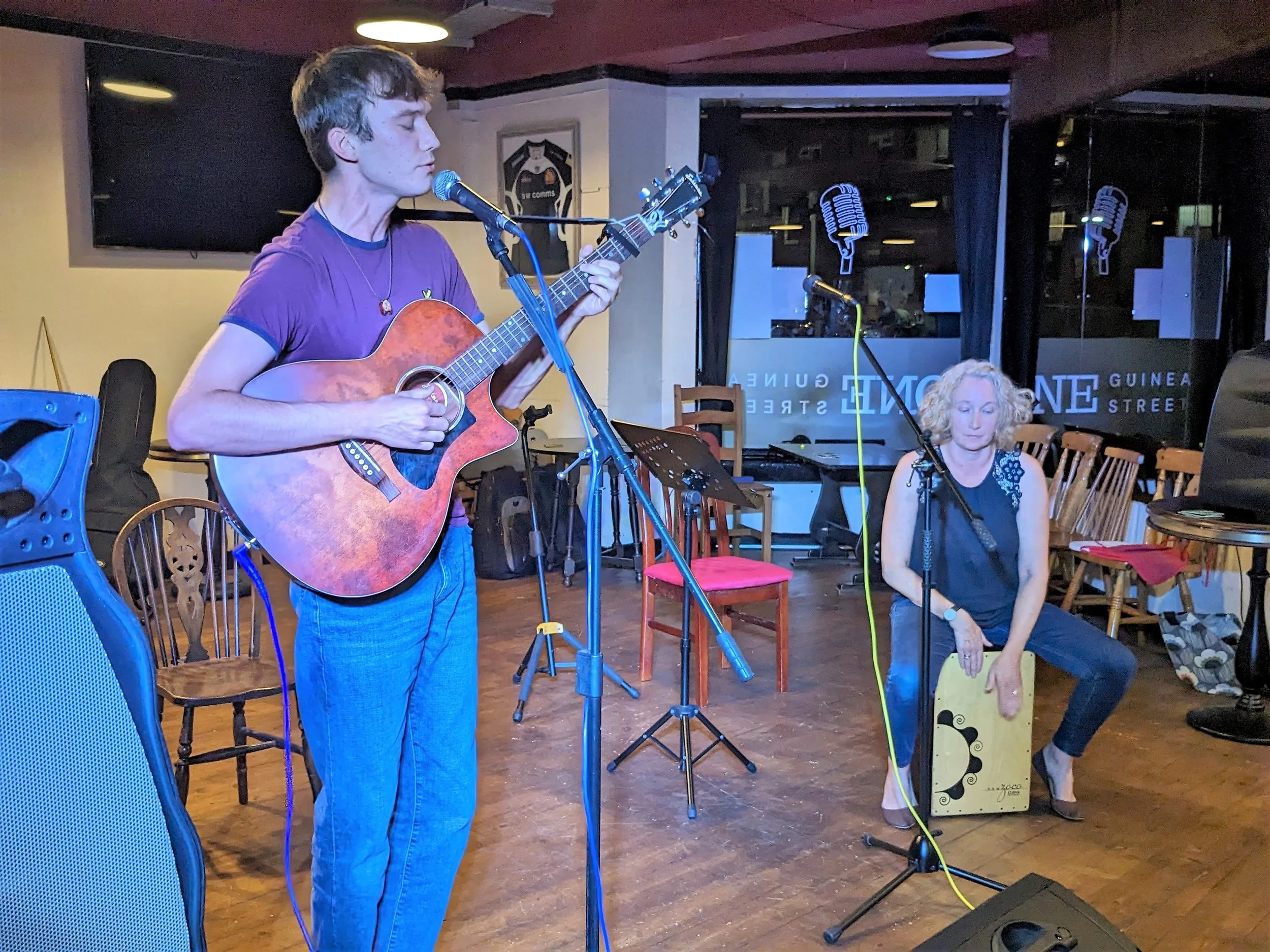A young man with curly hair playing an acoustic guitar near a microphone, with a woman sitting to his right, holding a percussion instrument, in a cozy indoor setting with chairs and music equipment.