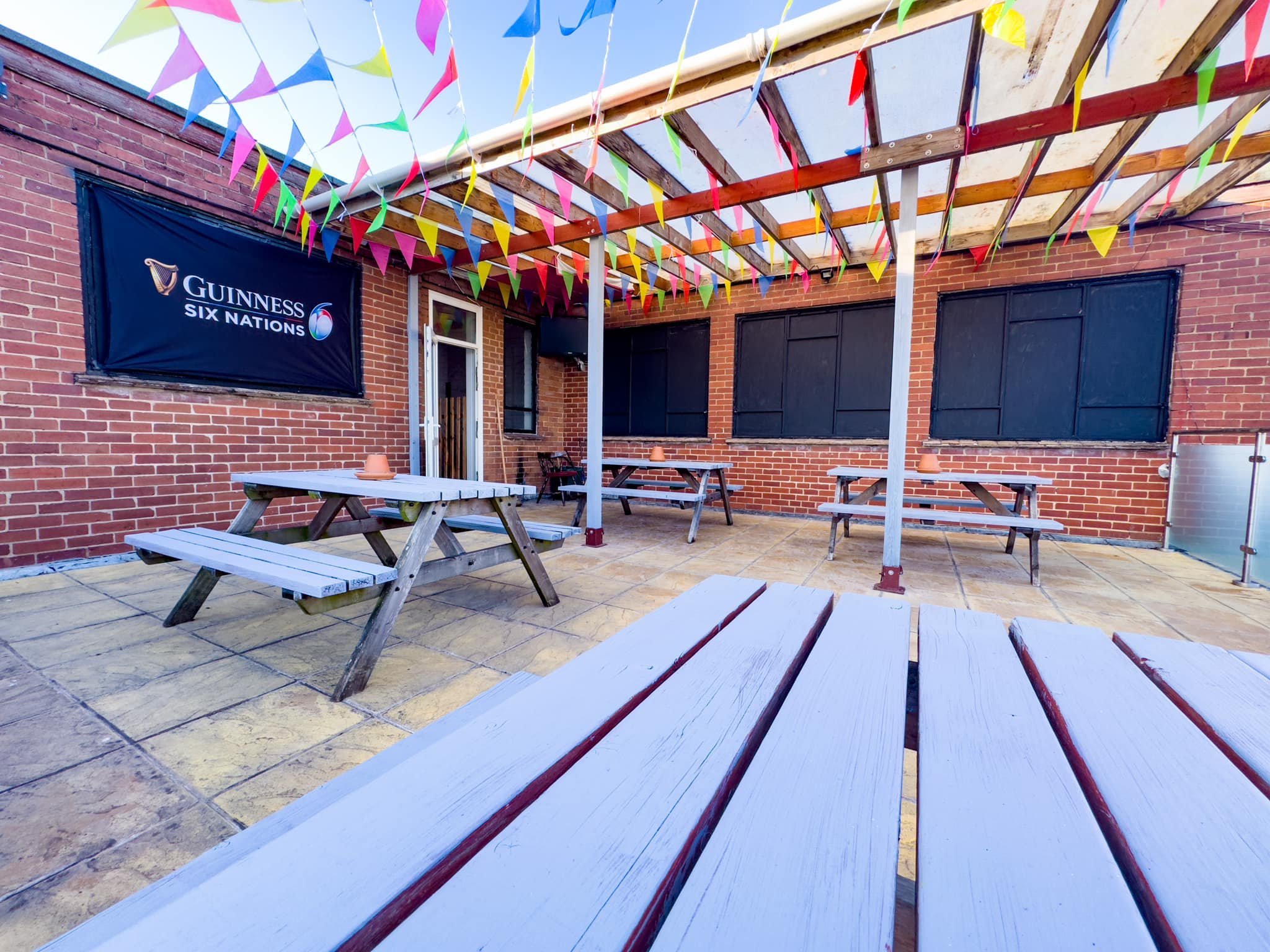 An outdoor patio area decorated with colorful pennant banners hanging from a pergola structure, with picnic tables and black window coverings on a brick building wall.