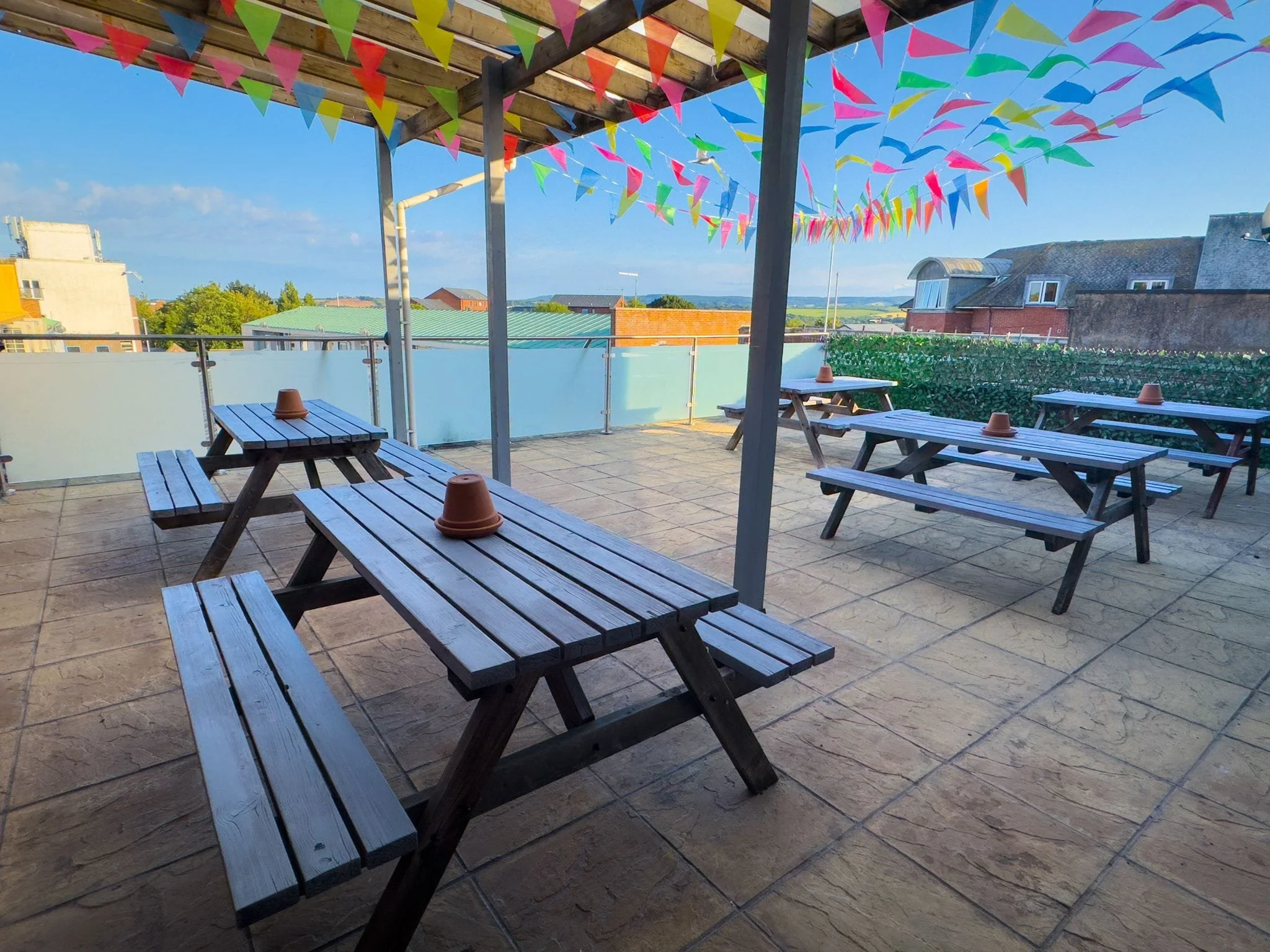 Outdoor patio area with wooden picnic tables, some with small terracotta plant pots on top. Overhead roof with multicolored triangular pennant banners strung across. Buildings and rooftops are visible in the background under a clear blue sky.