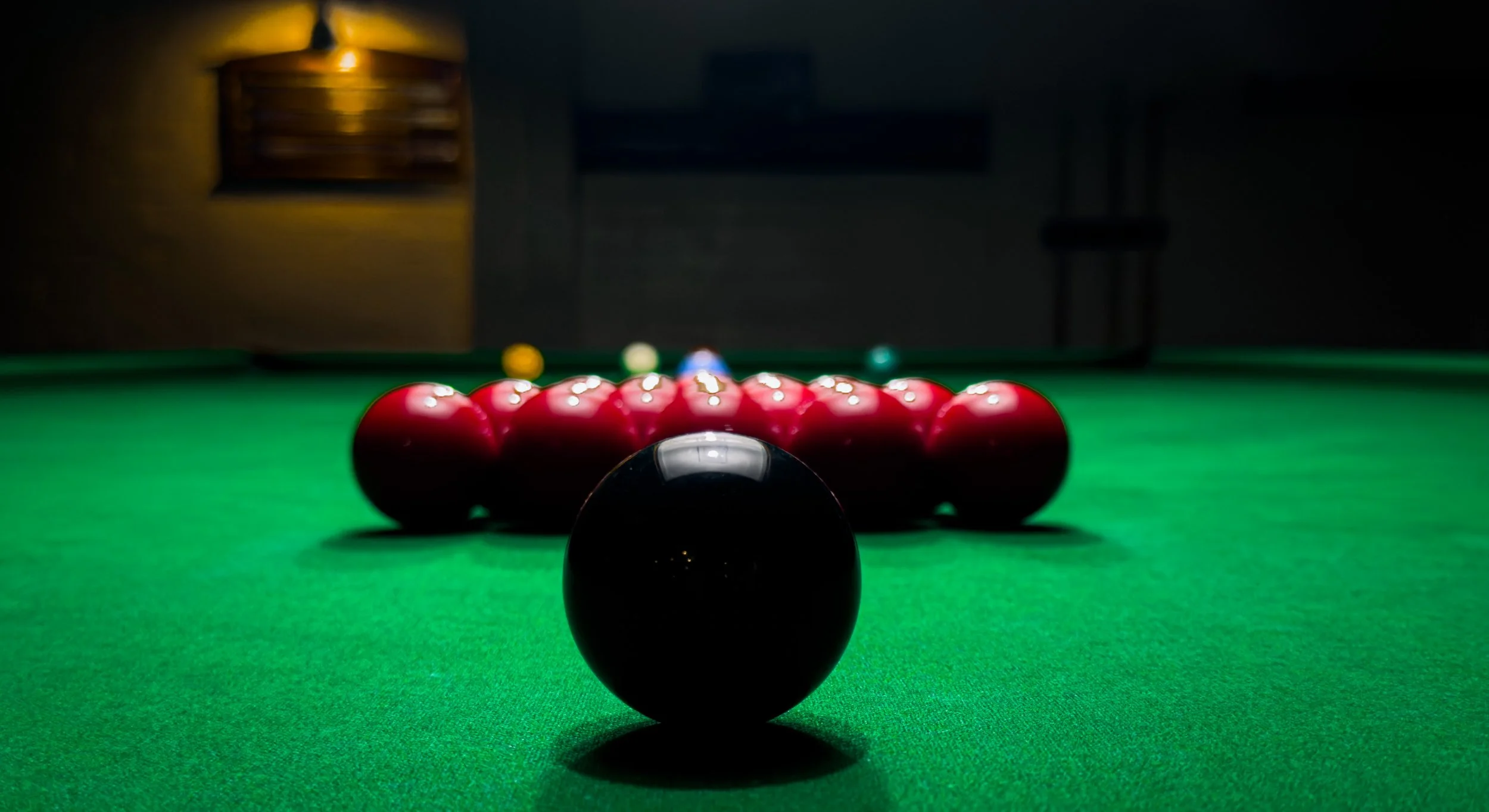 A snooker or pool table with a black cue ball in the foreground, surrounded by red balls arranged in a cluster, with colored balls in the background, in a dimly lit room.